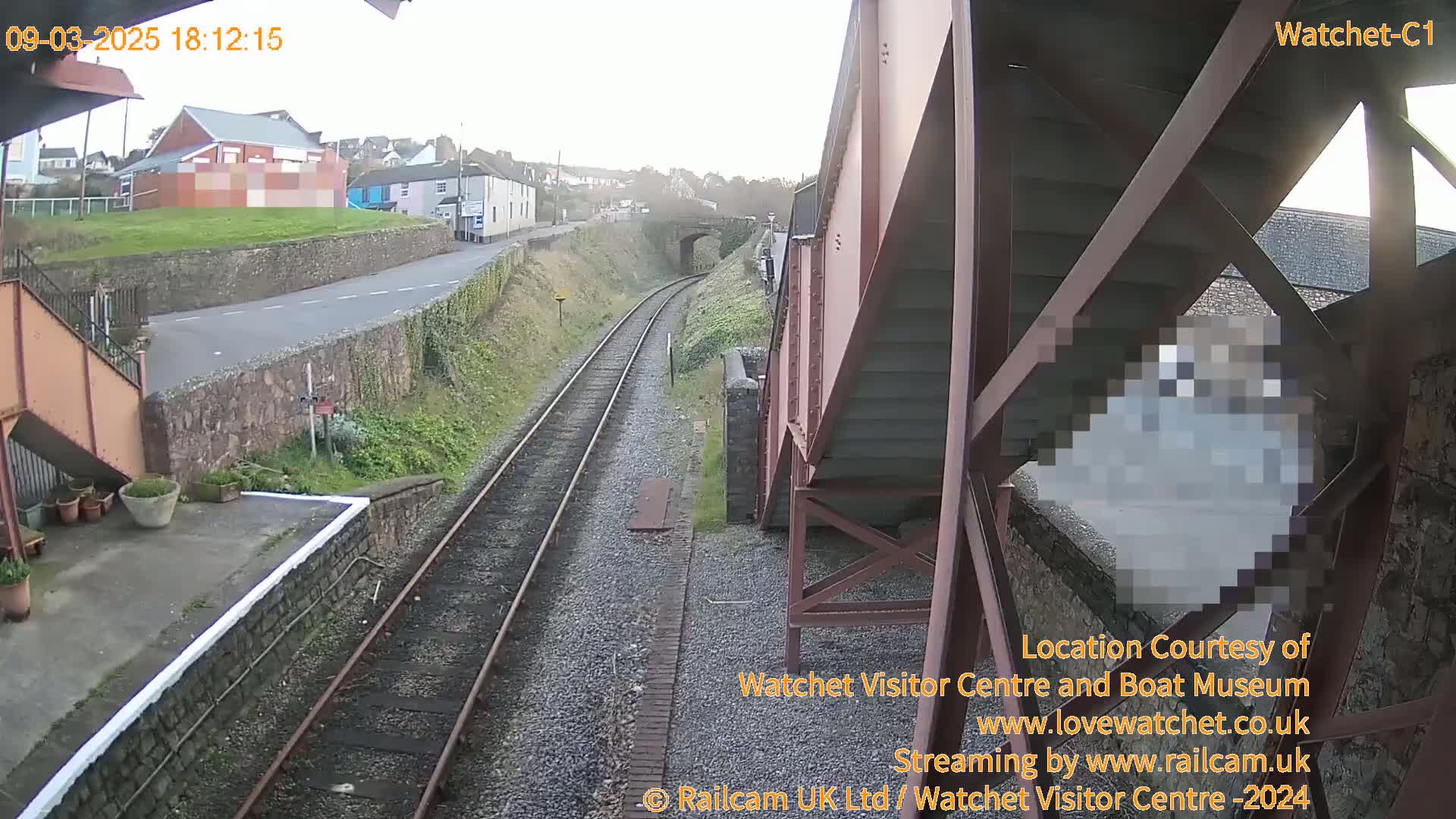 A view from a railway footbridge shows a railway line extending into the distance under an overcast sky, with a road and buildings visible beyond the tracks.