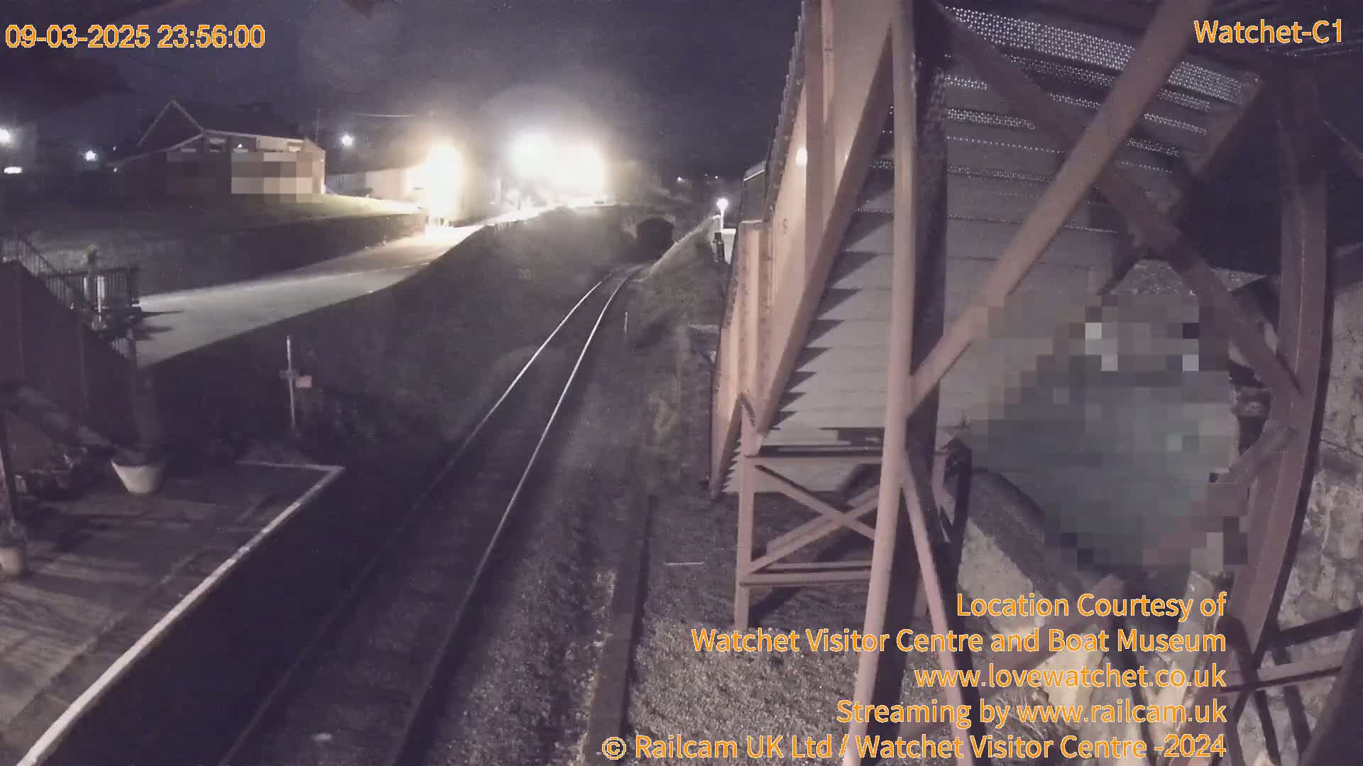 A nighttime view of a railway track and platform, with a metal structure overhead, under dark, possibly cloudy skies.