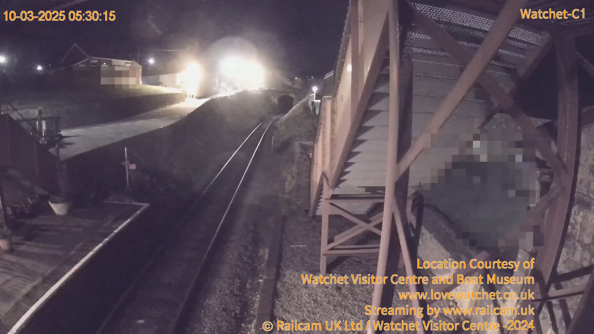 A nighttime view of a train passing by a railway station and a metal structure under a dark, clear sky.
