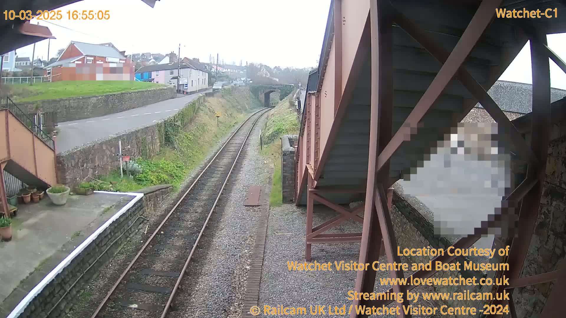 A view from a railway footbridge shows a cloudy day overlooking train tracks, a road, and buildings.
