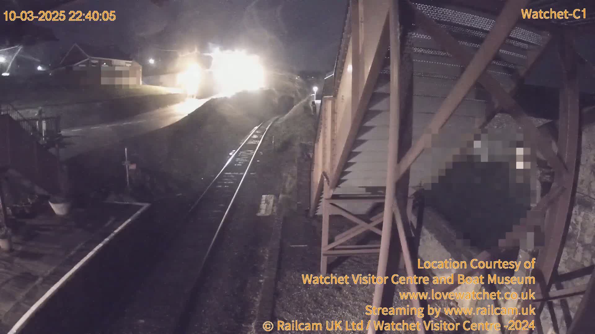 A brightly lit train approaches a railway station at night under dark, possibly overcast skies.