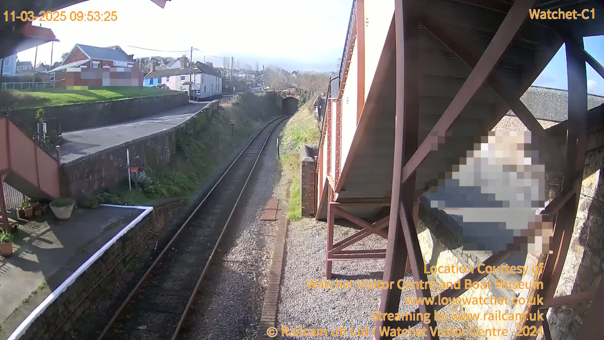 A sunny view from a railway bridge shows train tracks leading to a tunnel, with buildings and a road visible in the distance.