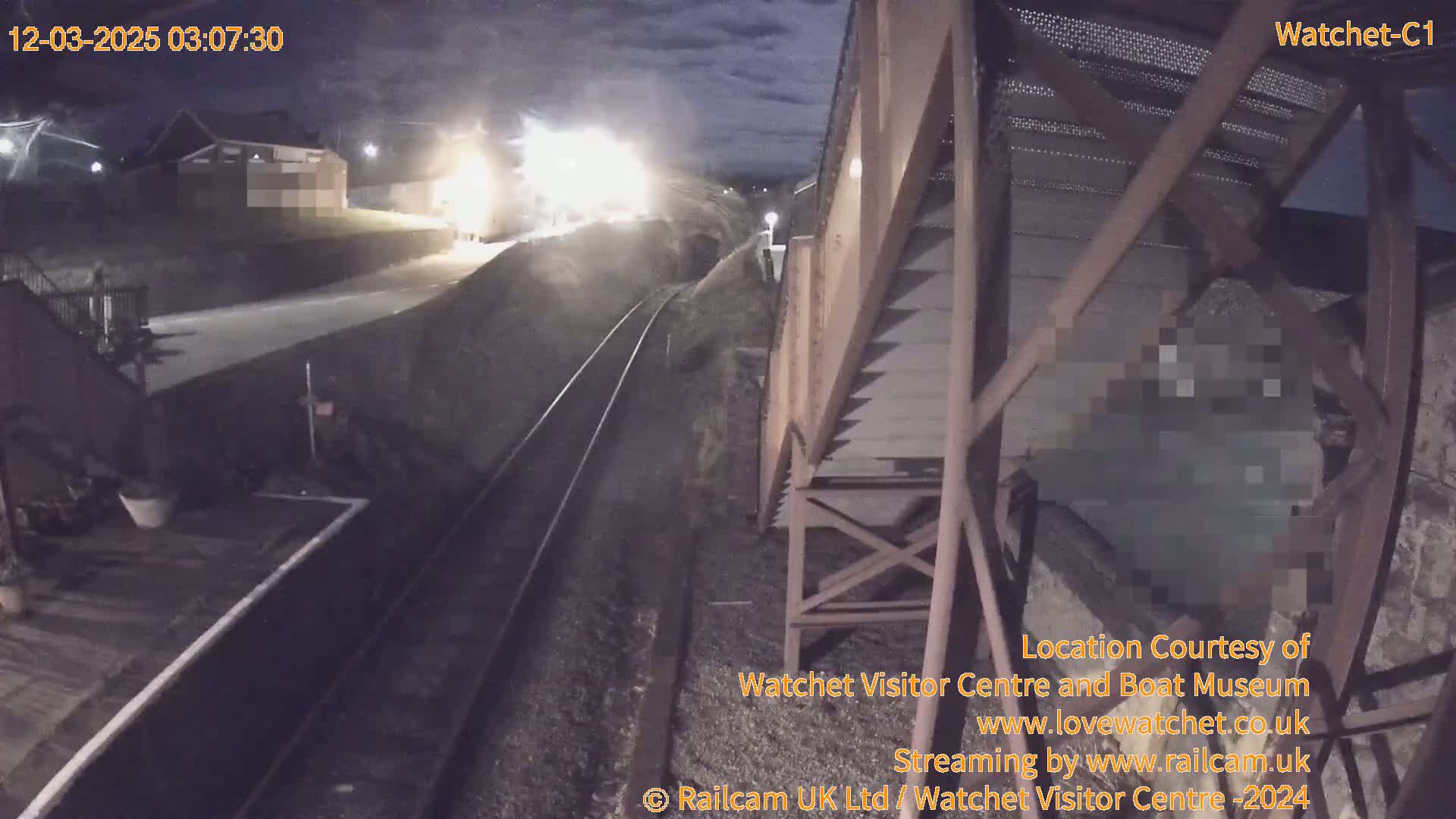 A nighttime view from a footbridge shows a train approaching a station platform under partly cloudy skies.