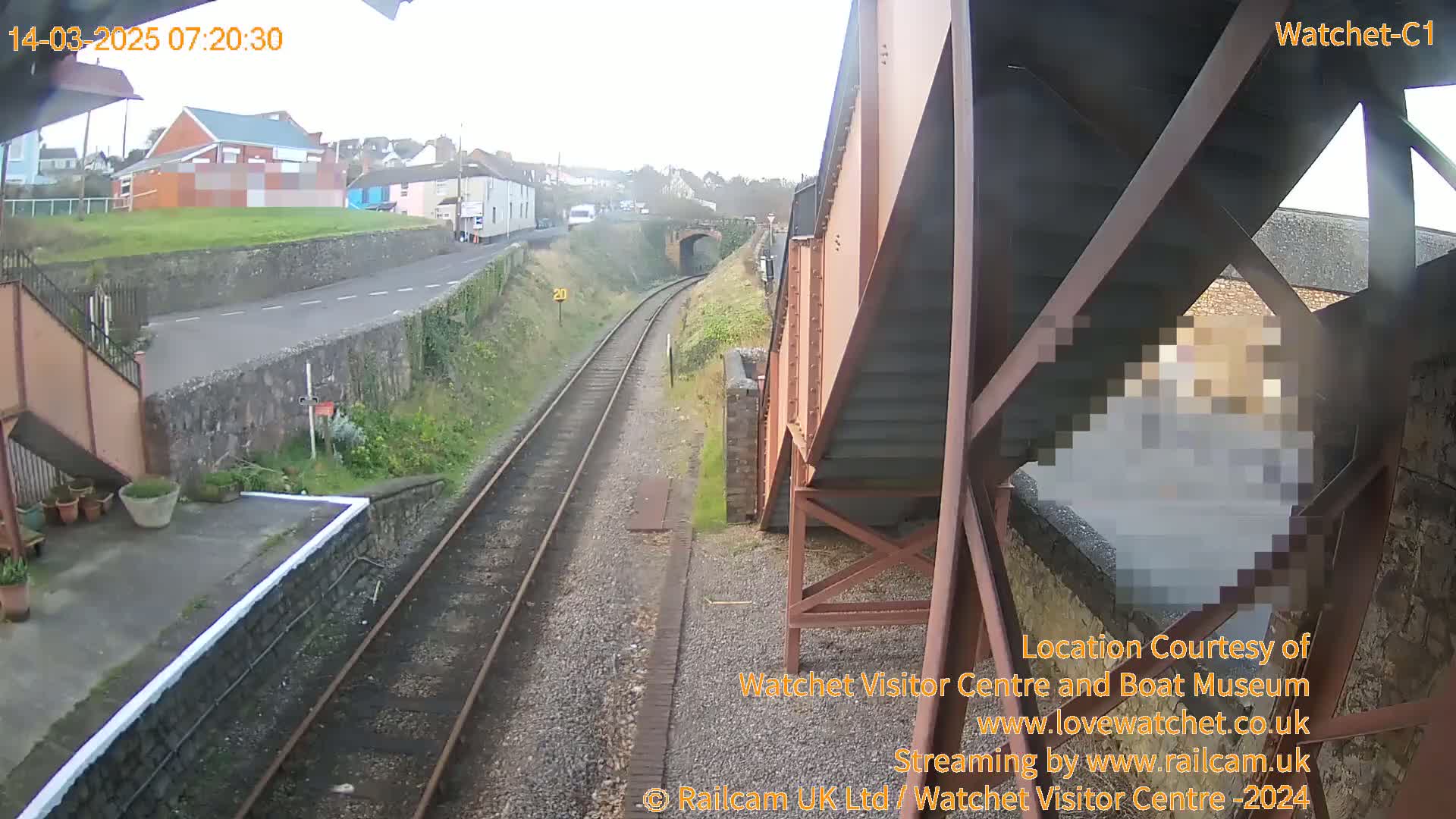 A view from a train platform shows railroad tracks extending into a tunnel under an overcast sky, with a road and buildings visible in the background.