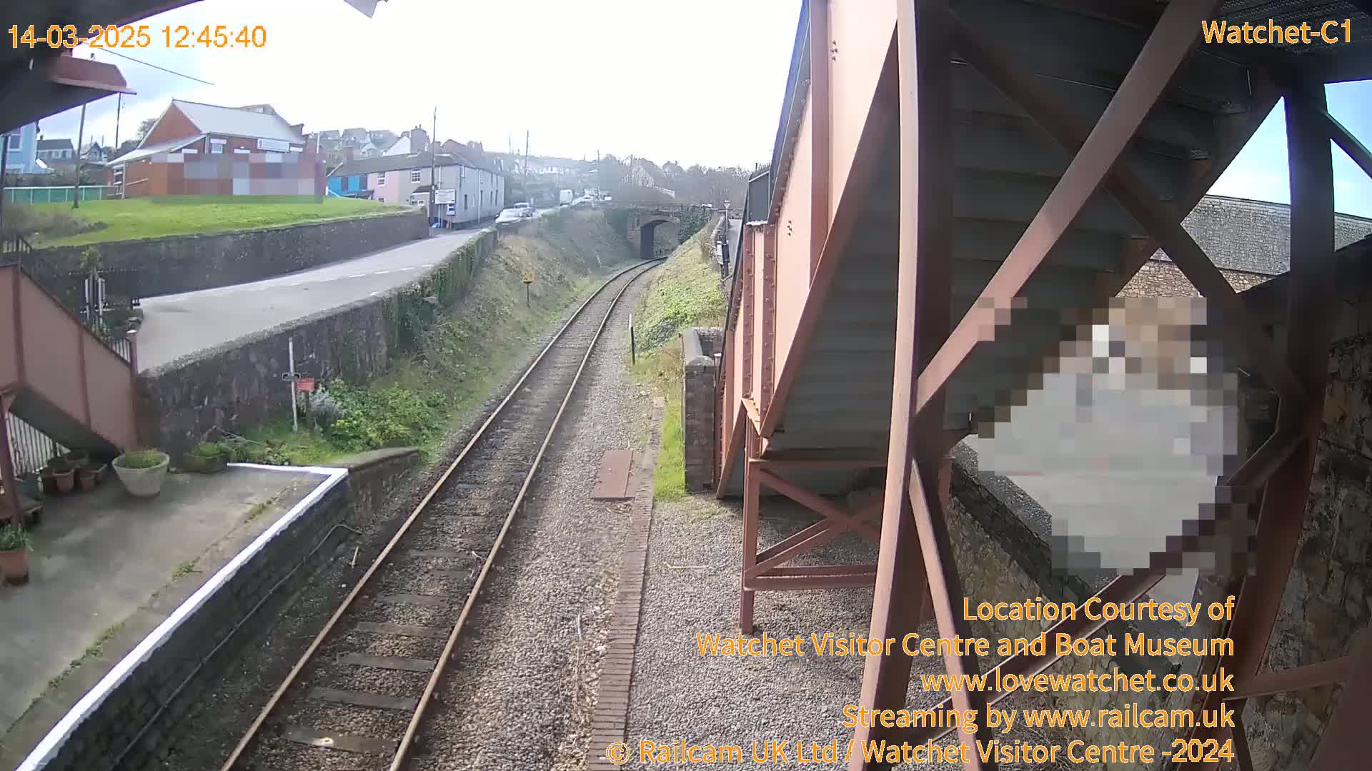 A view from a railway bridge on an overcast day shows train tracks extending into the distance, passing under a road bridge and beside a small town.
