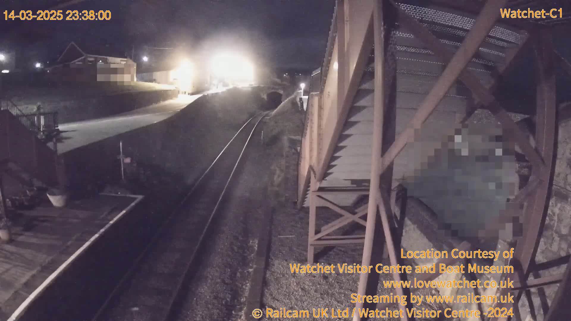 A nighttime, overhead view of a train track near a building and a metal structure, under dark skies.