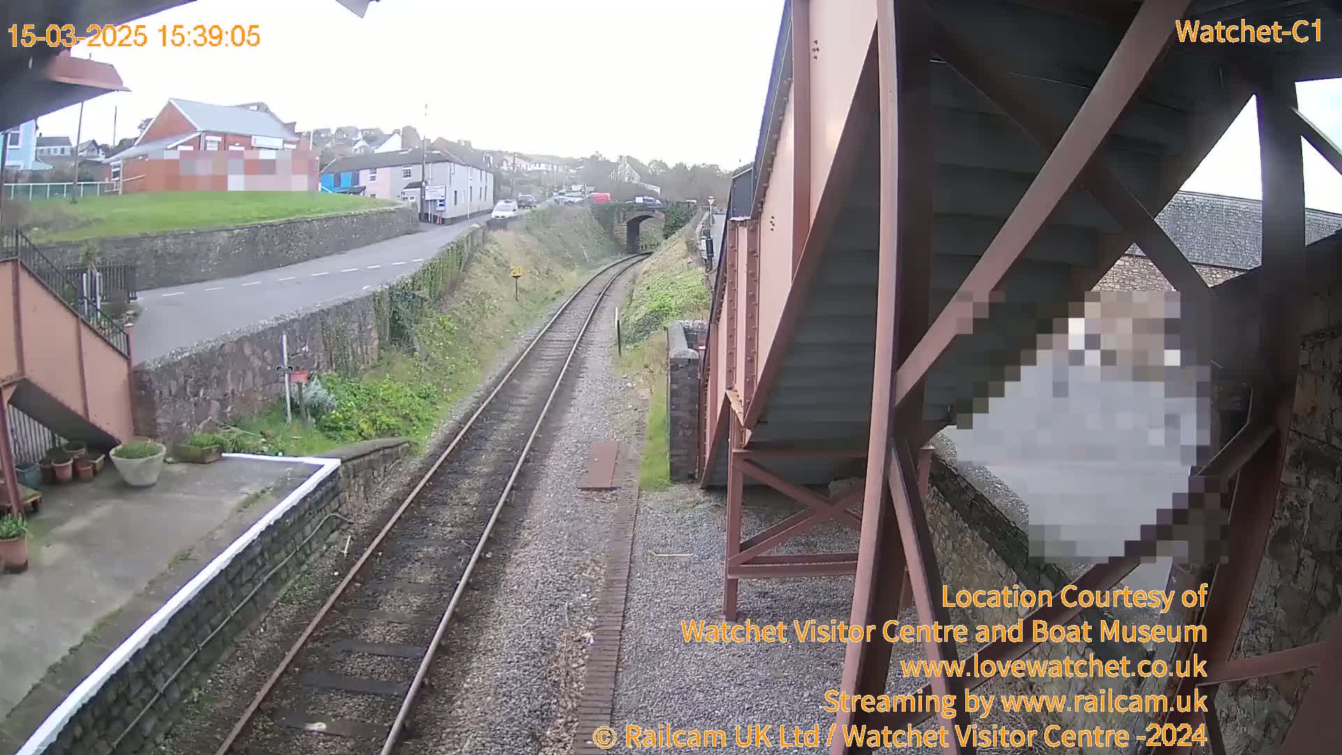 A view from under a railway bridge shows a train track leading toward a small town under an overcast sky.