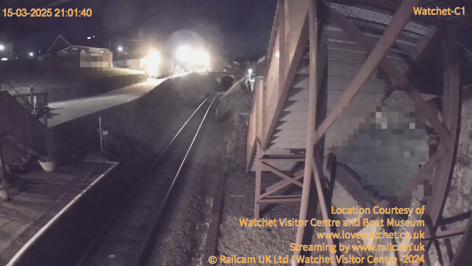 A nighttime view from a railway bridge shows train tracks leading to a tunnel, with a building and some lights visible in the distance.