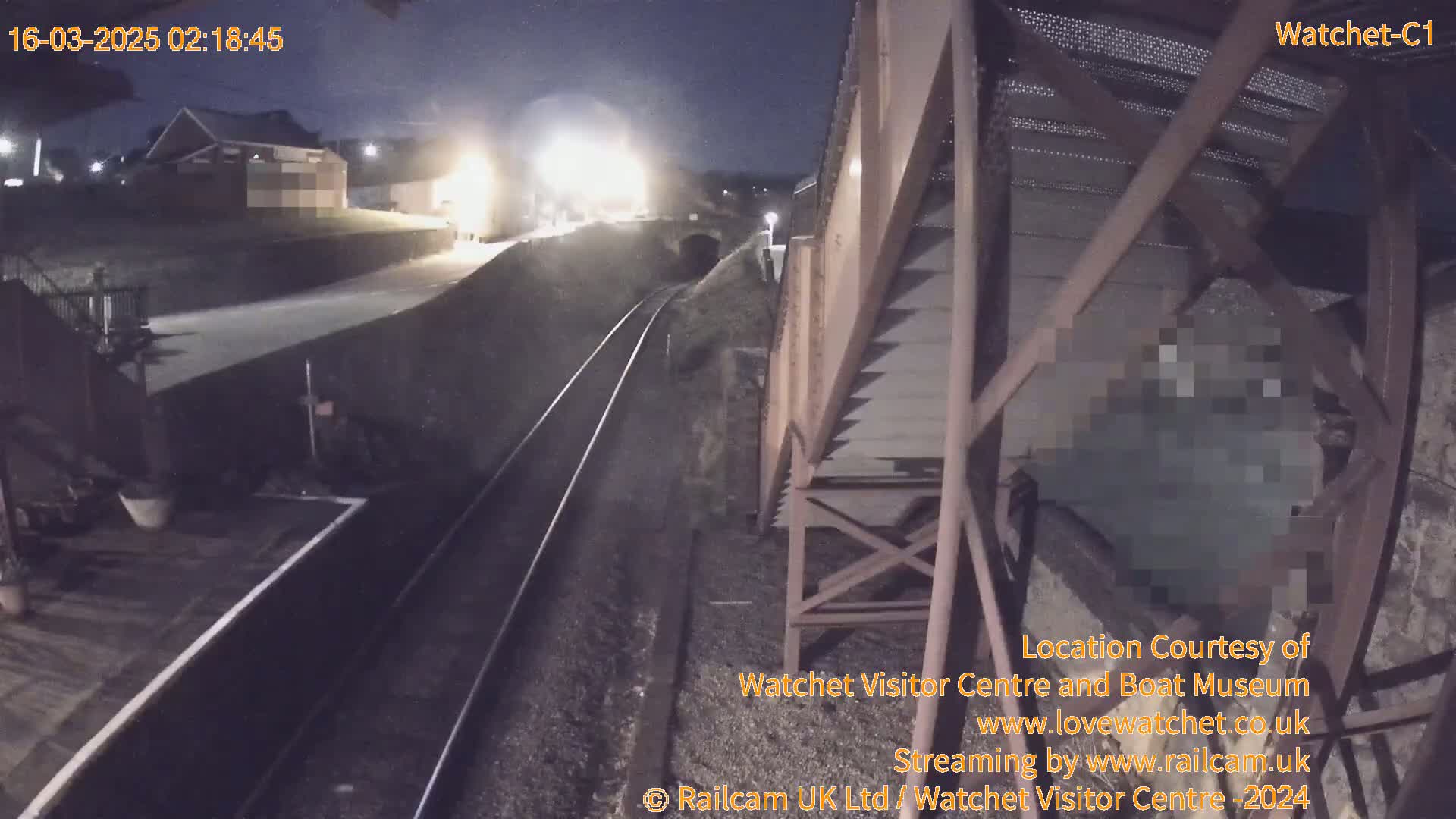 A nighttime view from a railway bridge shows train tracks, a dimly lit building, and a train approaching a tunnel.