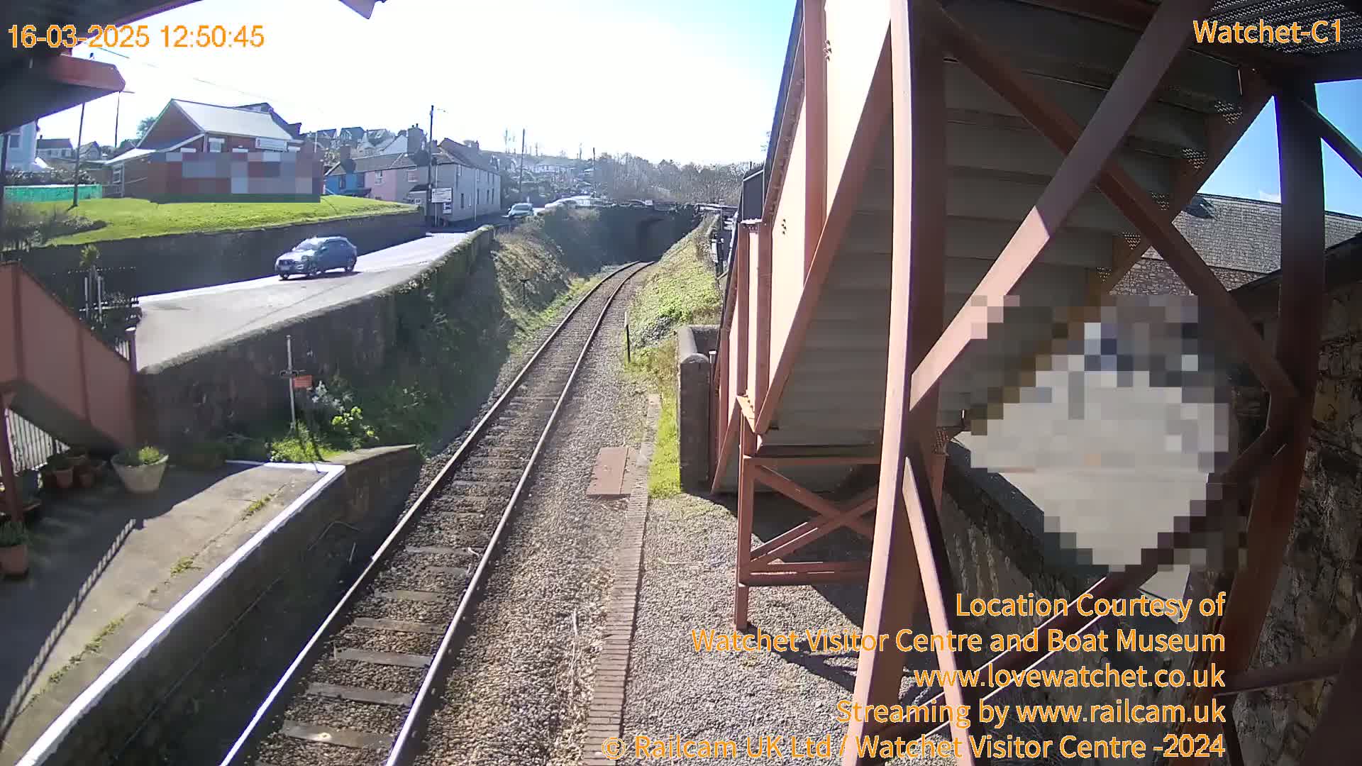 A sunny view from a railway bridge shows a single car on a road above train tracks, leading to a tunnel under a small town.