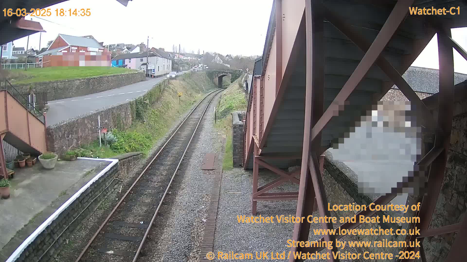 A view of a railway track and a road from an overhead metal structure on an overcast day, with a person partially obscured by the structure.