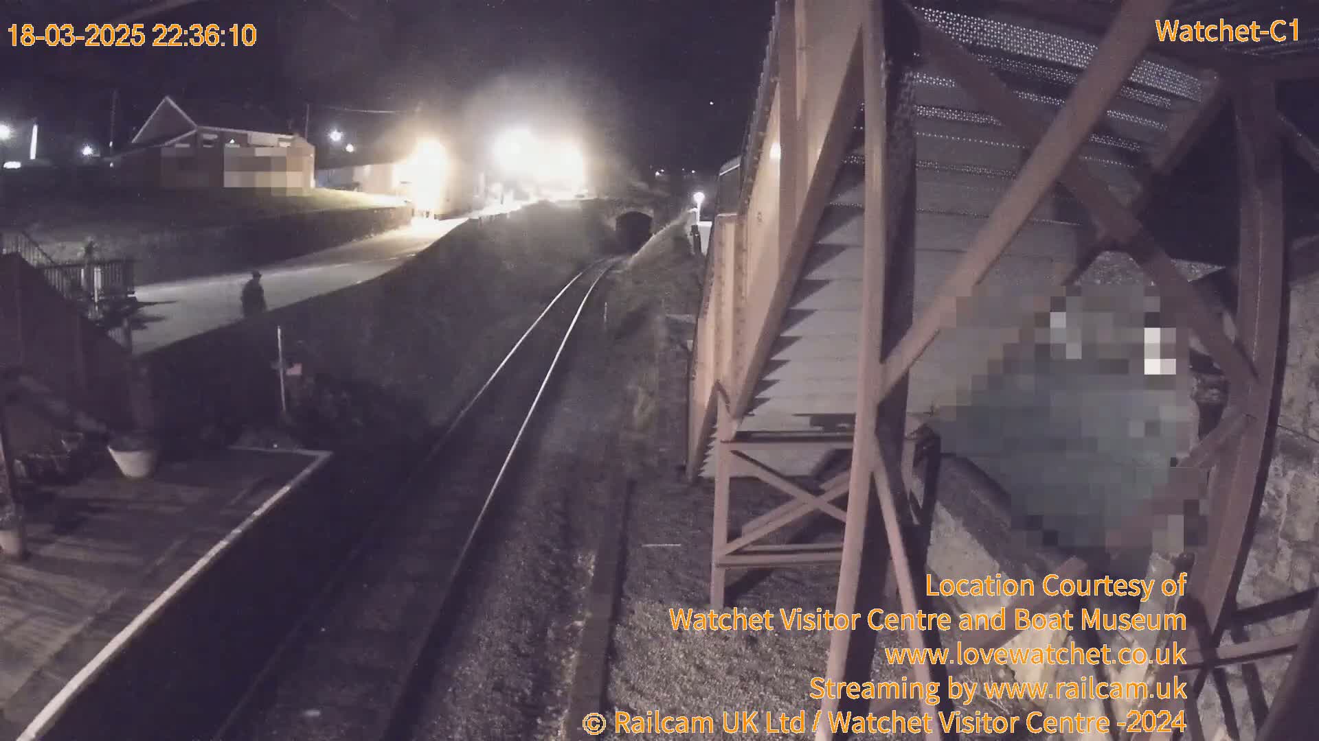 A nighttime view of a railway track and a metal structure, with a person visible in the distance and a building illuminated in the background.