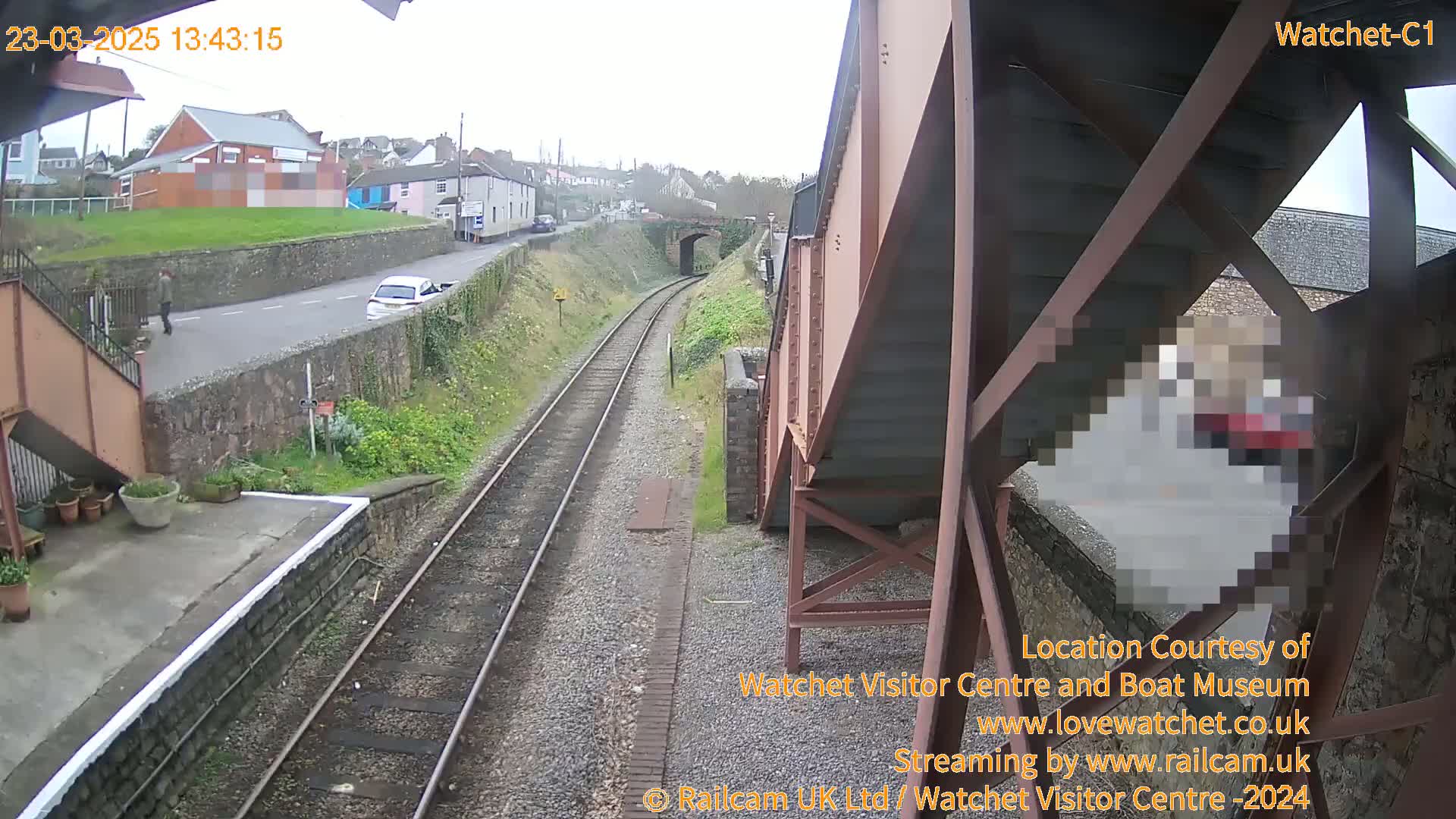 A view from an elevated railway bridge shows a train track, a road with a car and pedestrian, and overcast weather.
