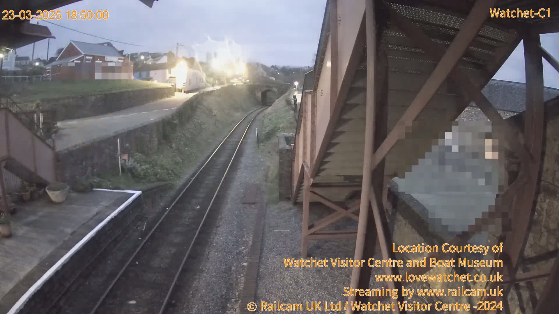 A view of a train station platform and tracks at dusk under overcast skies, with a building visible in the distance.