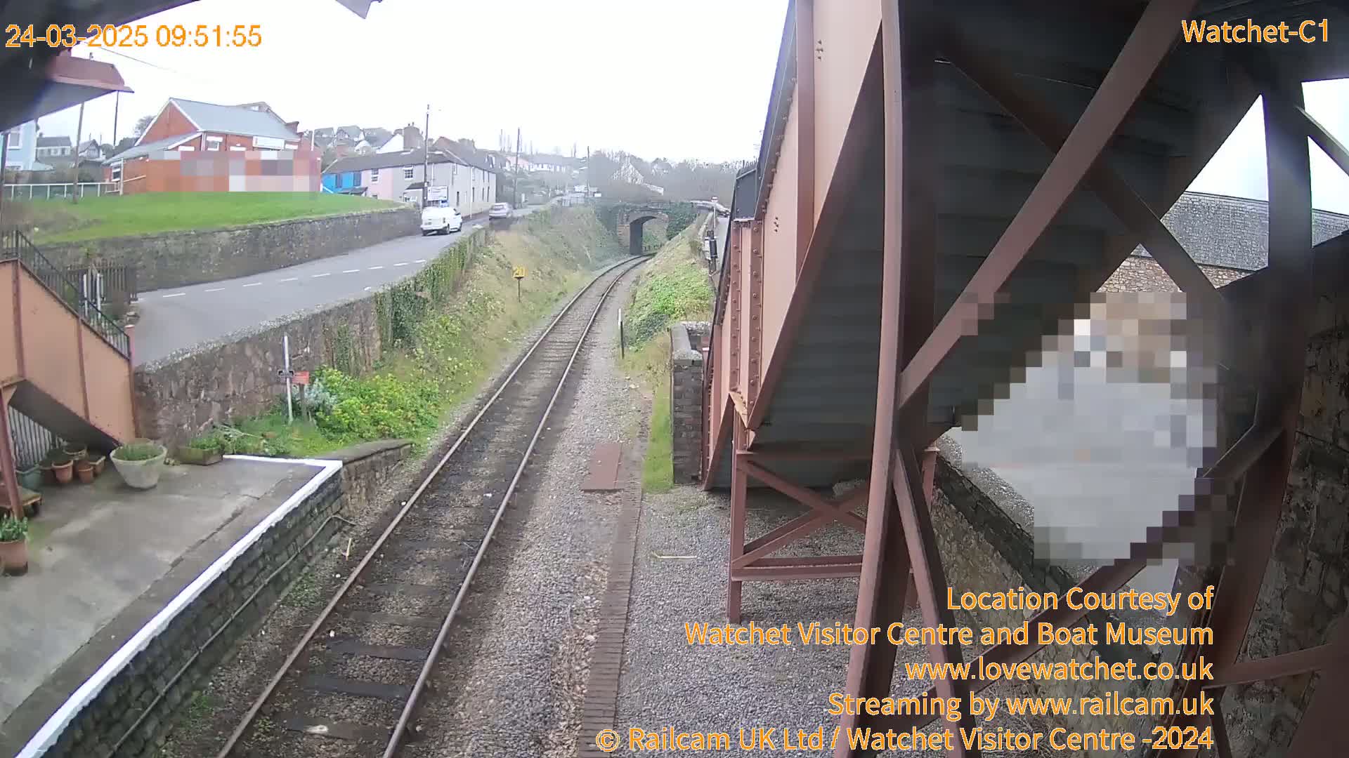 A view from under a metal footbridge shows a railway line and a road next to a small town on an overcast day.