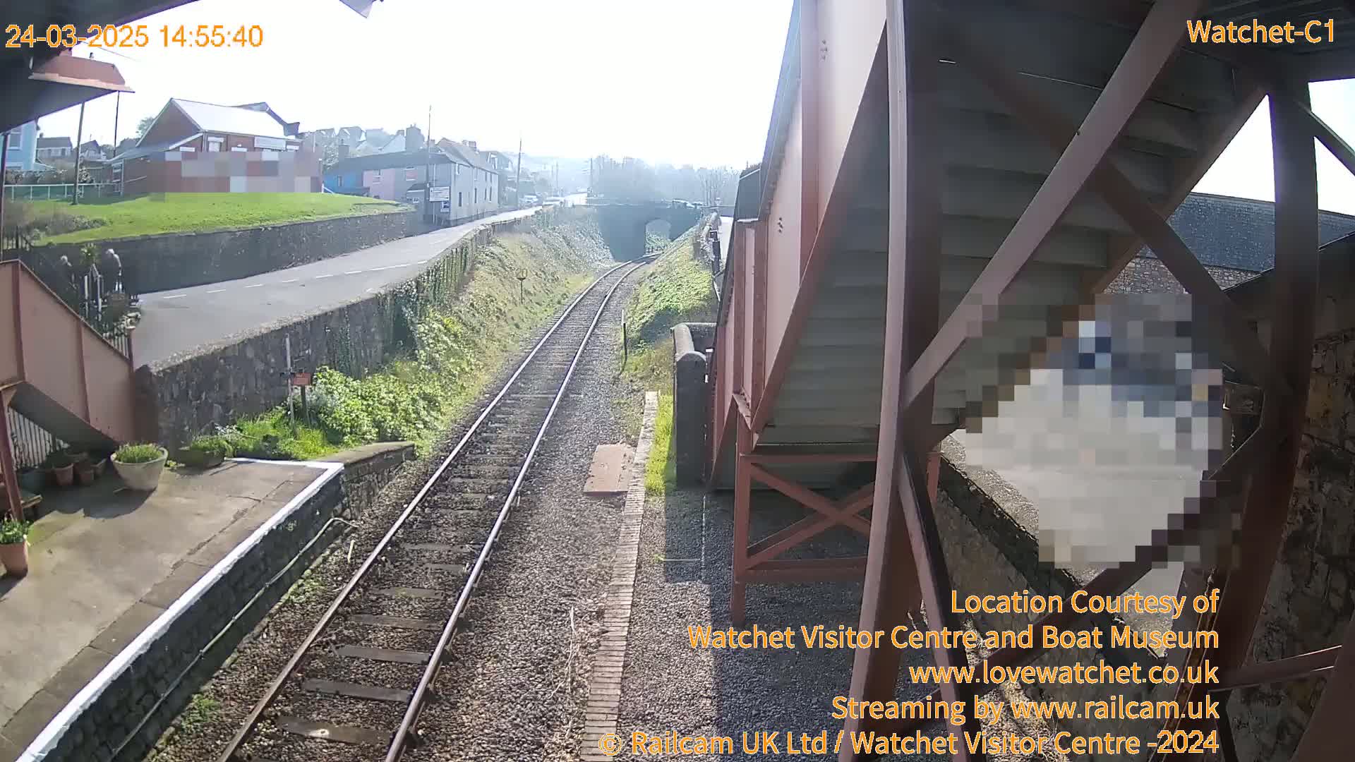 A sunny view of a railway track and station platform, with a road and buildings visible in the distance.