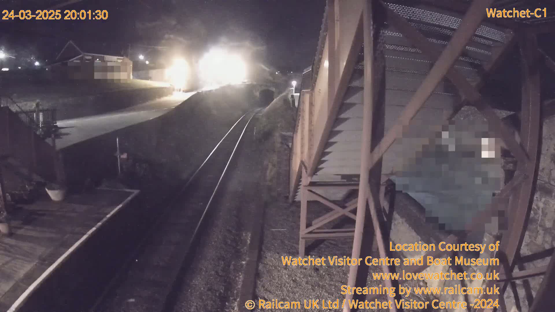 A nighttime view from a bridge overlooking a train track, with a train approaching a tunnel under the bridge.