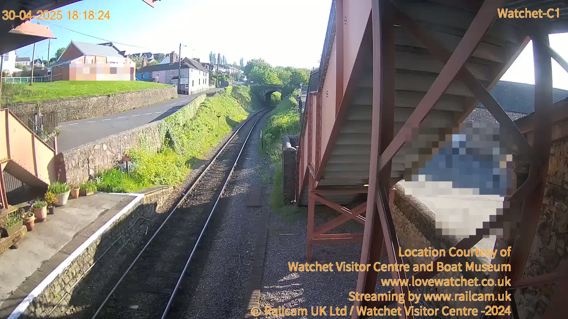 A sunny view from a railway footbridge shows train tracks extending into the distance, passing by a small town with buildings and a grassy area.