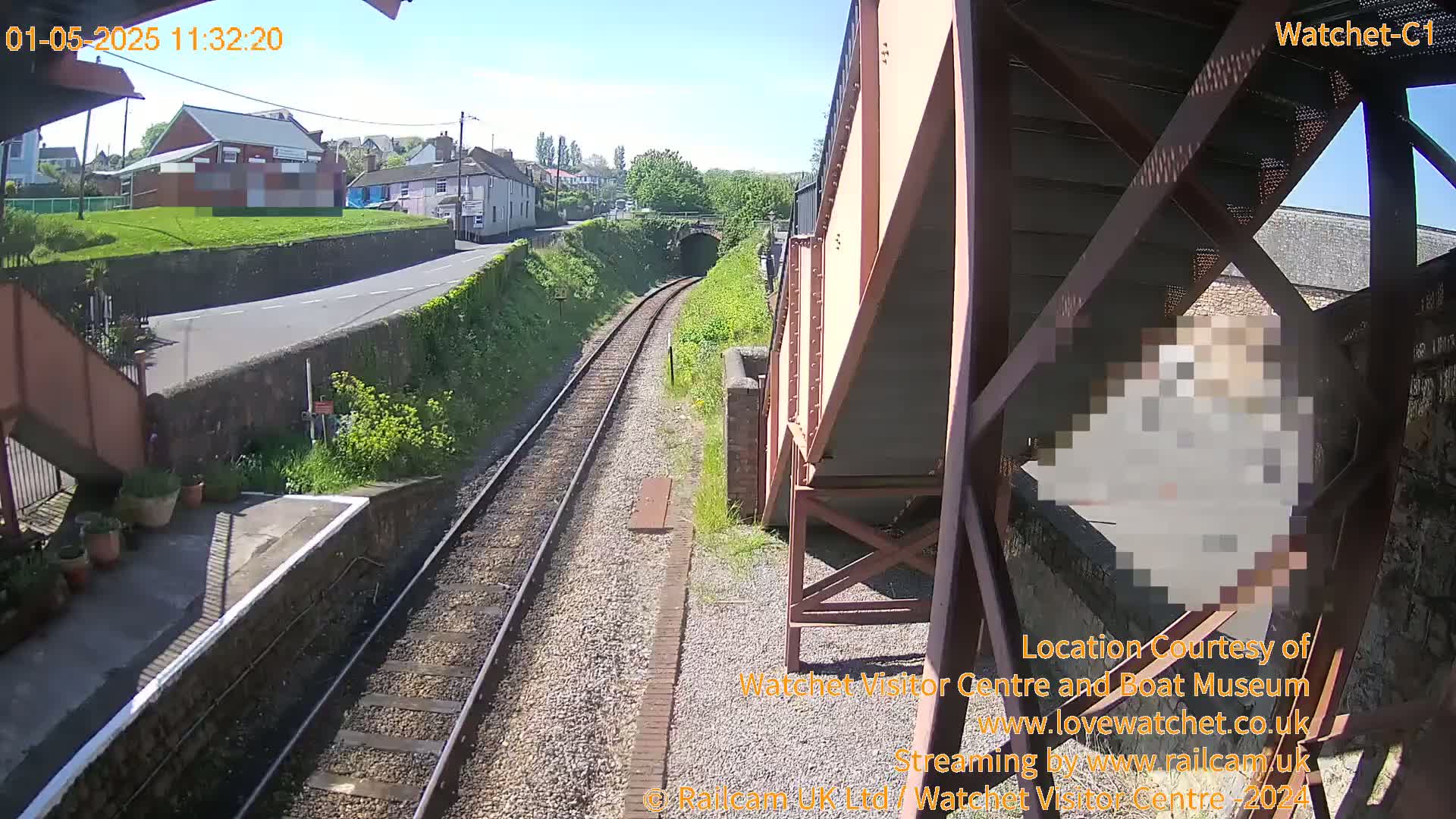 A sunny view of a railway track extending toward a tunnel, with a road and buildings visible in the background.
