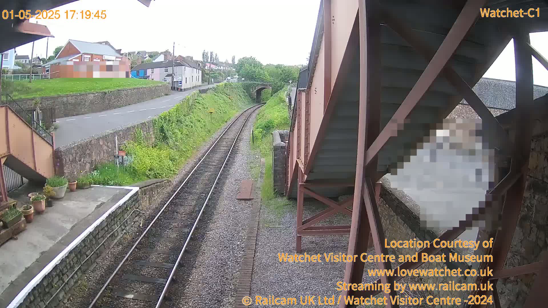 A view from a railway bridge shows train tracks leading toward a tunnel under a road, with buildings and green vegetation visible in the background under an overcast sky.