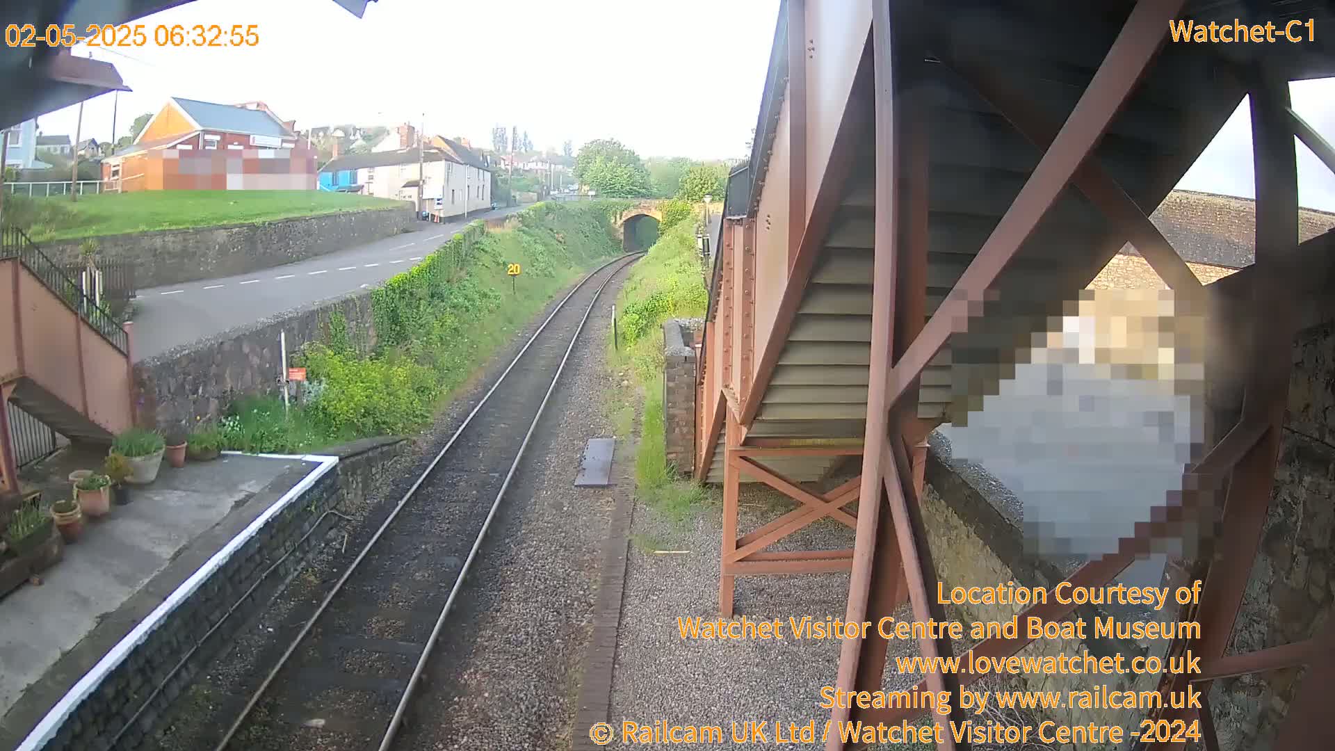 A view from a railway bridge on a partly sunny morning shows train tracks leading toward a tunnel, with buildings and a road visible in the distance.