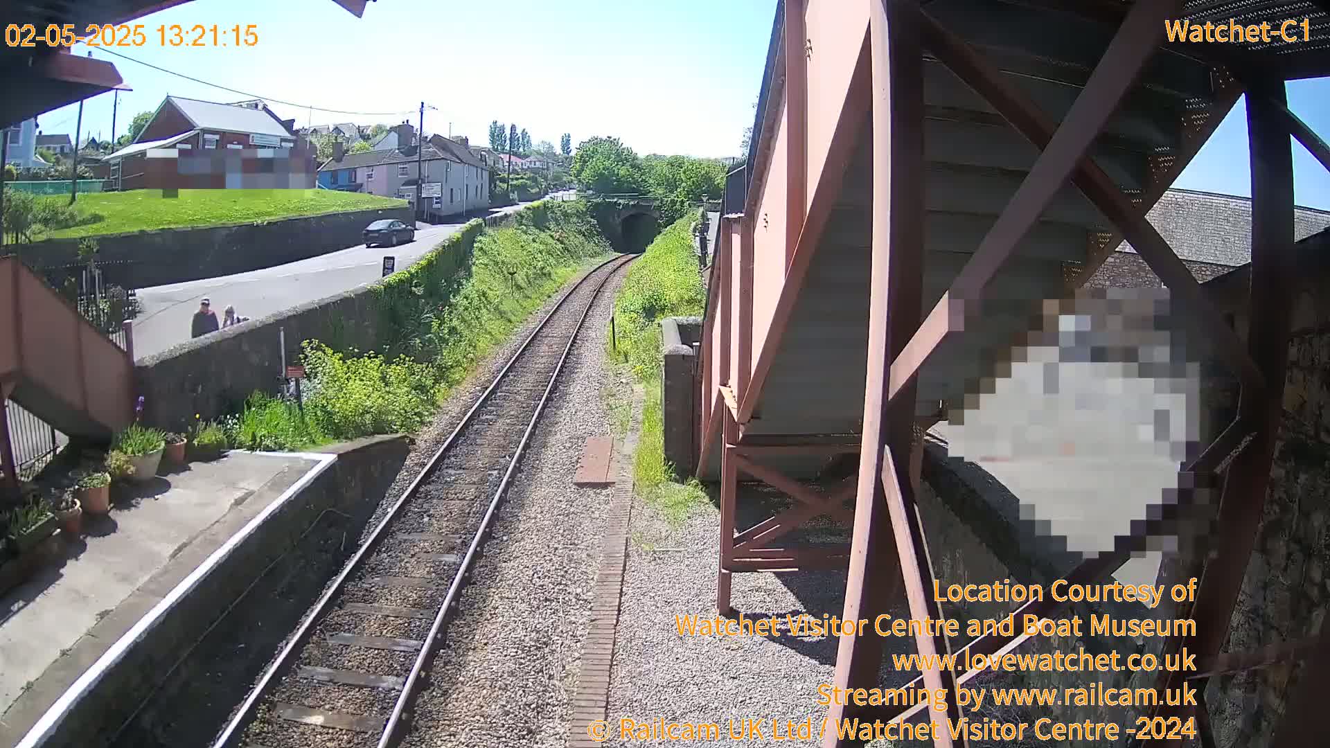 A sunny view from a train platform shows a railway track leading to a tunnel, a road with a car and pedestrians, and buildings in the background.
