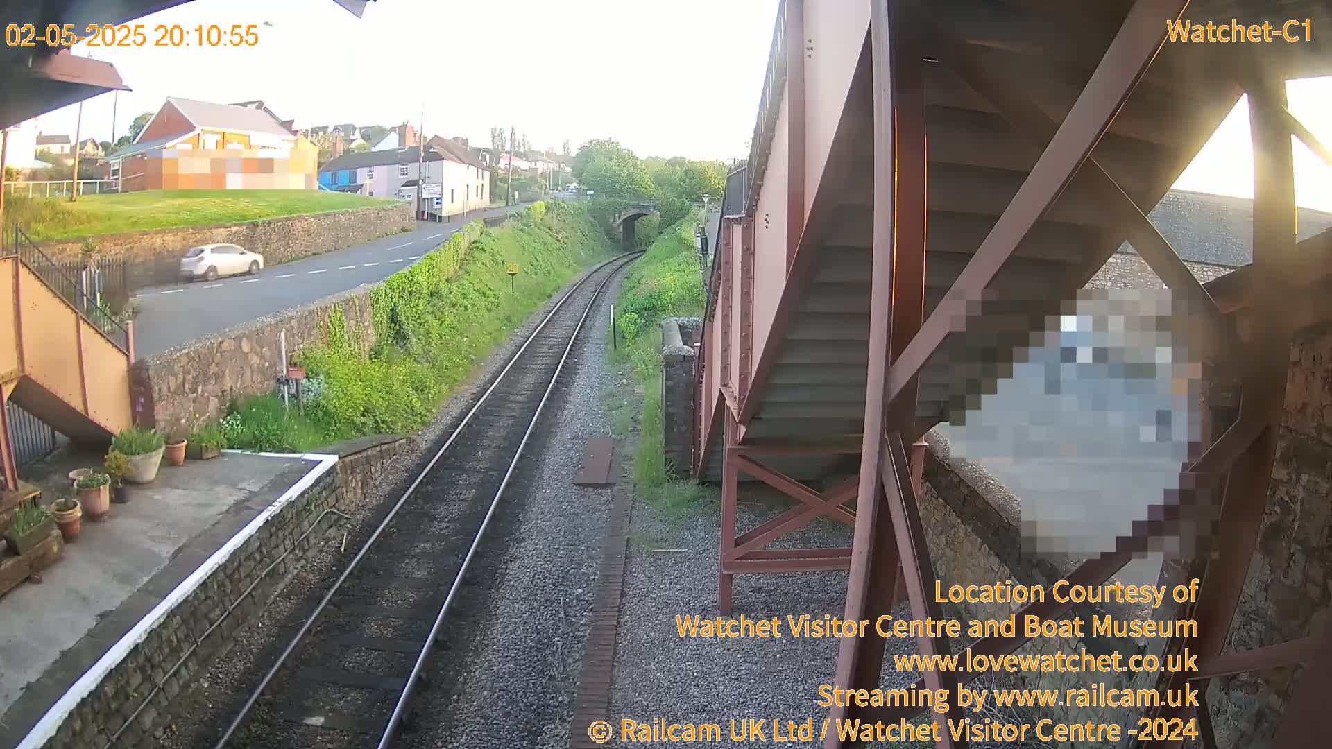A view from a footbridge shows train tracks and a road with a car, under a clear evening sky.