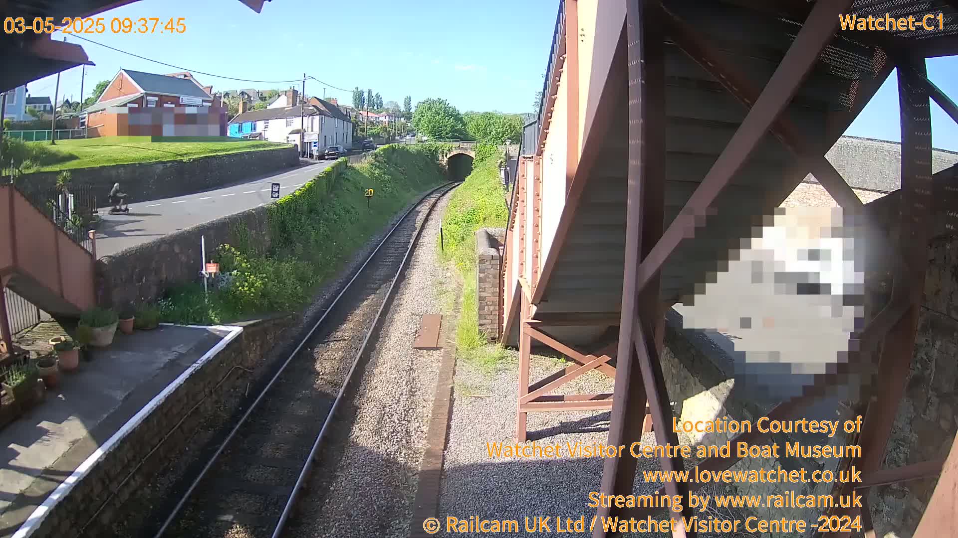 A sunny view from a railway footbridge shows train tracks leading to a tunnel, a person on a scooter on a road above, and buildings in the background.