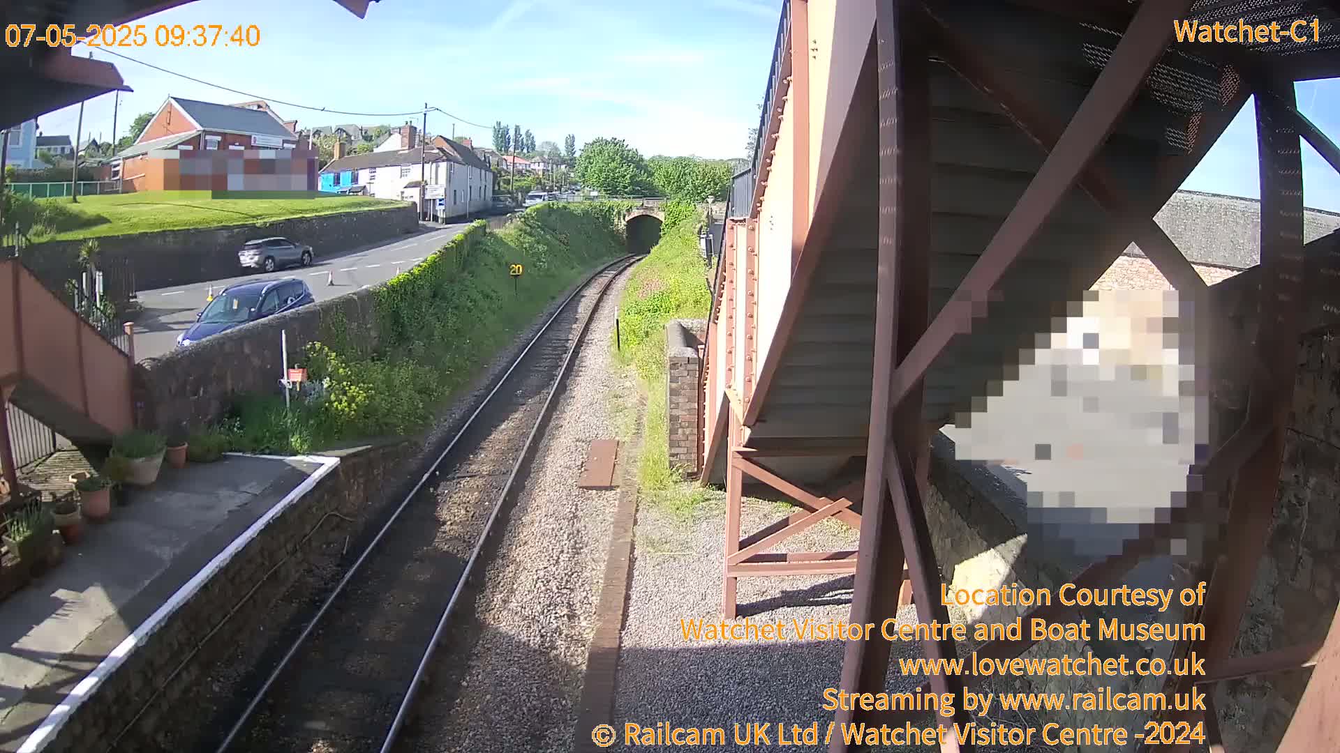 A high-angle view of a train track leading towards a tunnel, with cars parked on a road next to a grassy embankment under a sunny sky.