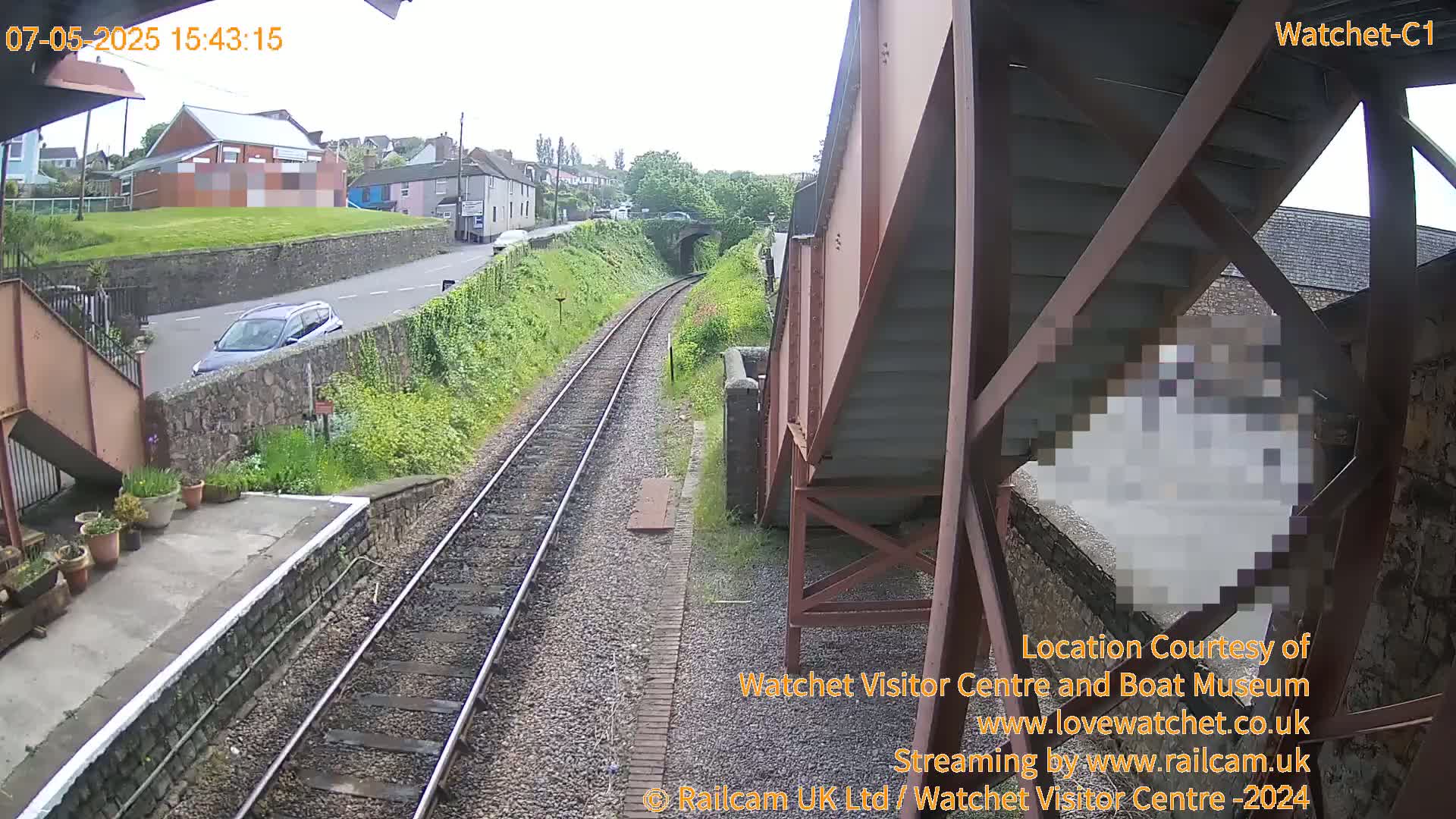 A view from a train platform on a partly cloudy day shows train tracks leading to a tunnel, a road with a car, and buildings in the distance.