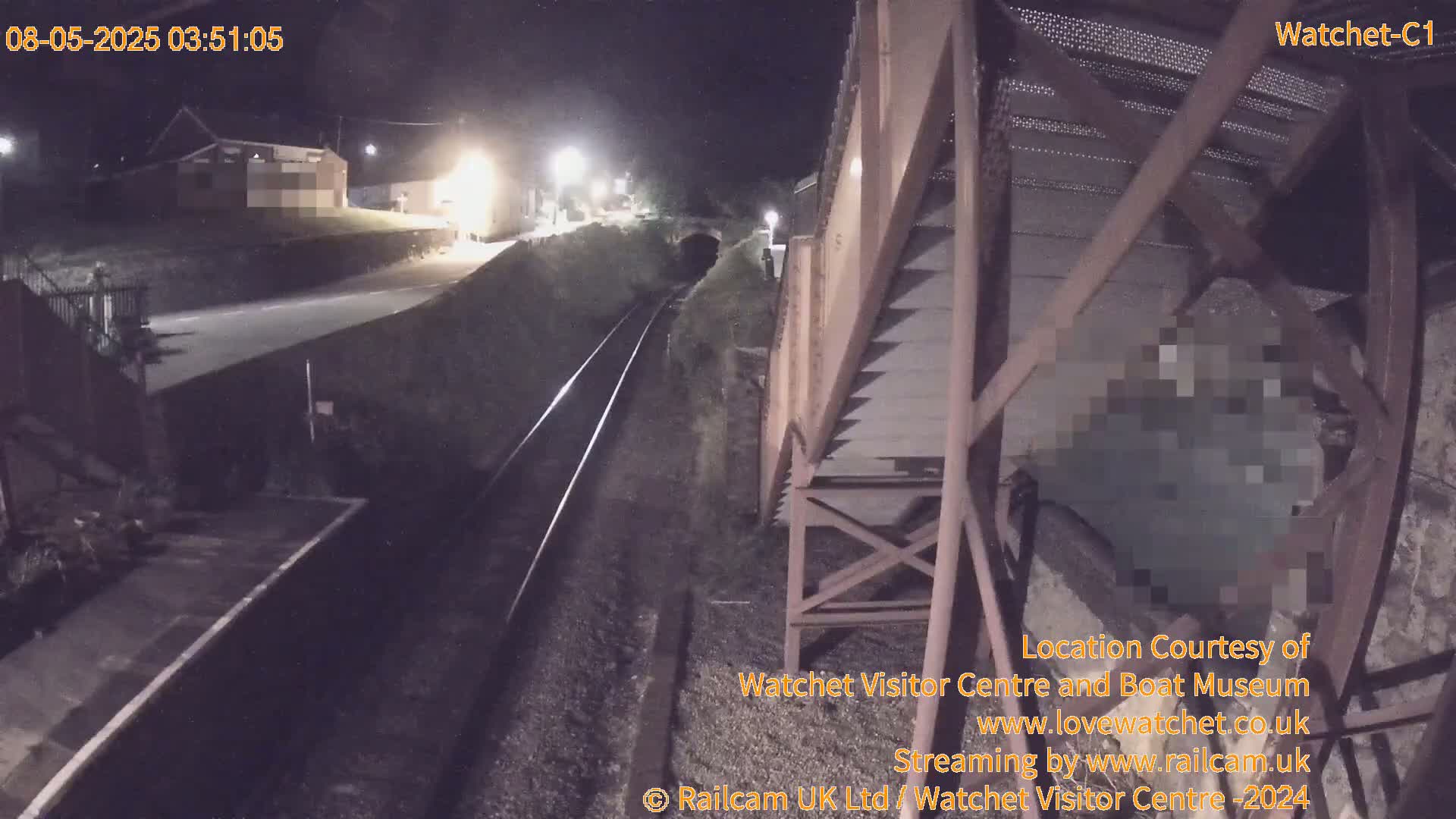 A nighttime view from a bridge overlooking a railway track, with a building and road visible in the distance under dark, clear skies.