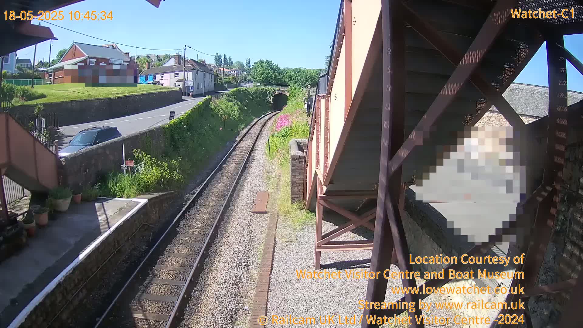 A sunny view of a railway track leading toward a tunnel, with a road and buildings visible in the distance, and a partially visible person obscured by pixels in the foreground.