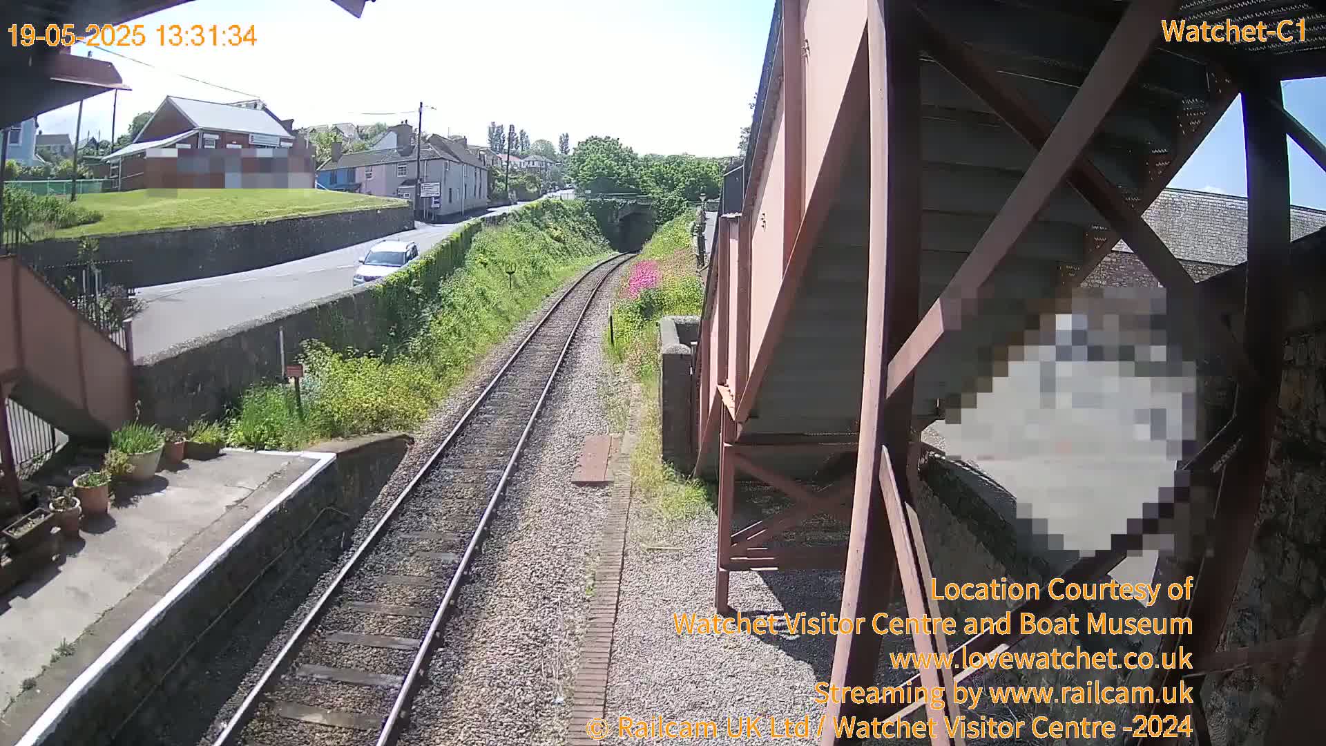 A sunny view from a railway platform shows a road, houses, and railway tracks leading to a tunnel, with a car visible on the road.
