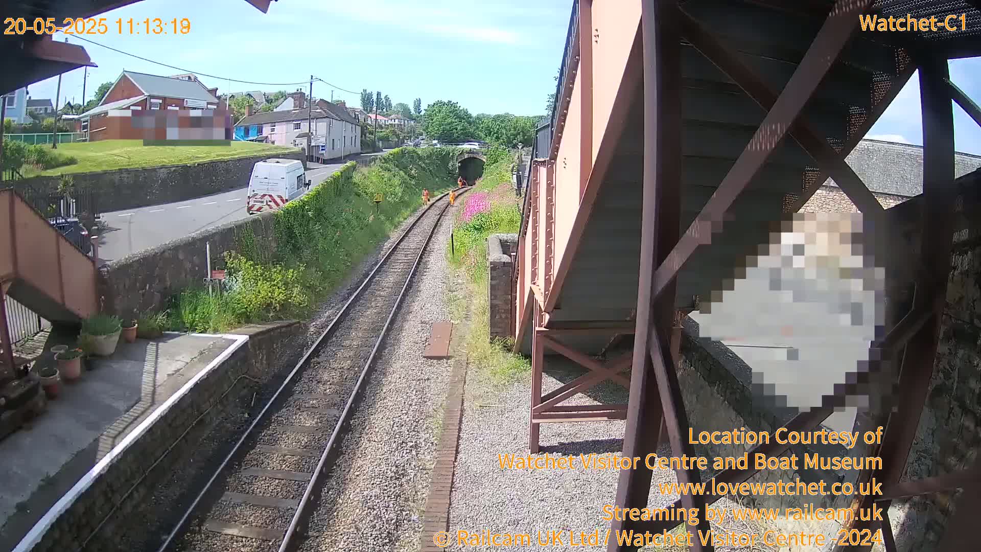 A sunny view from a railway bridge shows train tracks leading to a tunnel, with workers in orange, a white van on a road, and buildings in the distance.