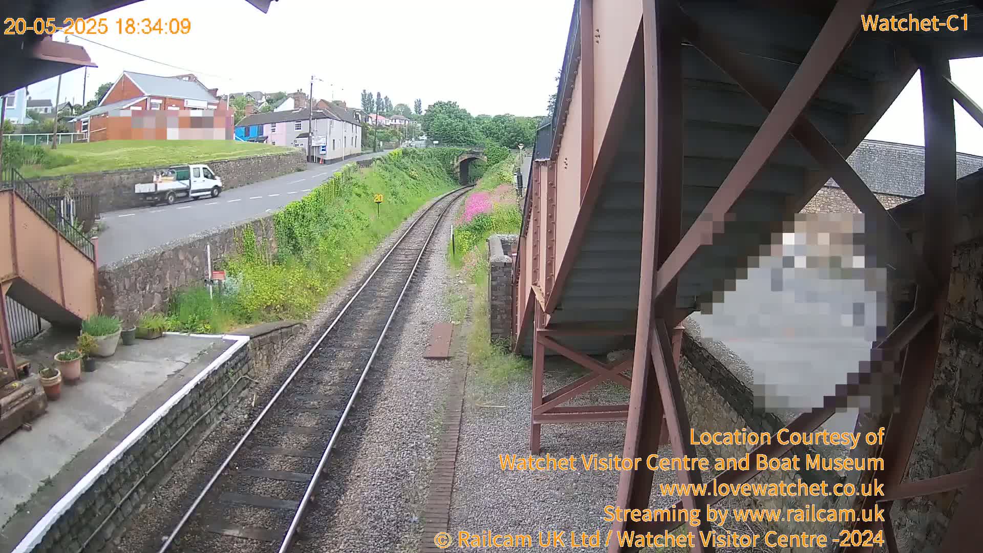A view from a railway bridge on an overcast day shows train tracks, a small white truck on a road, and some buildings in the distance.