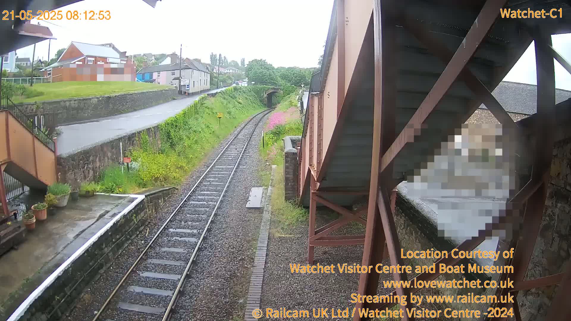A rain-slicked railway track leads toward a tunnel, passing beside a small station with buildings and greenery visible in the background.
