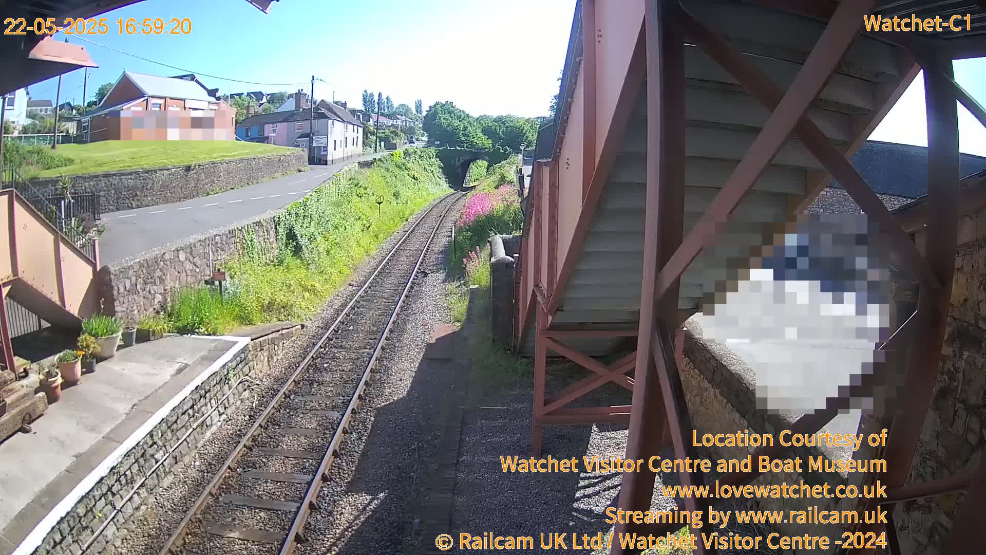 A sunny view of train tracks, a station platform, and nearby buildings from an elevated vantage point.