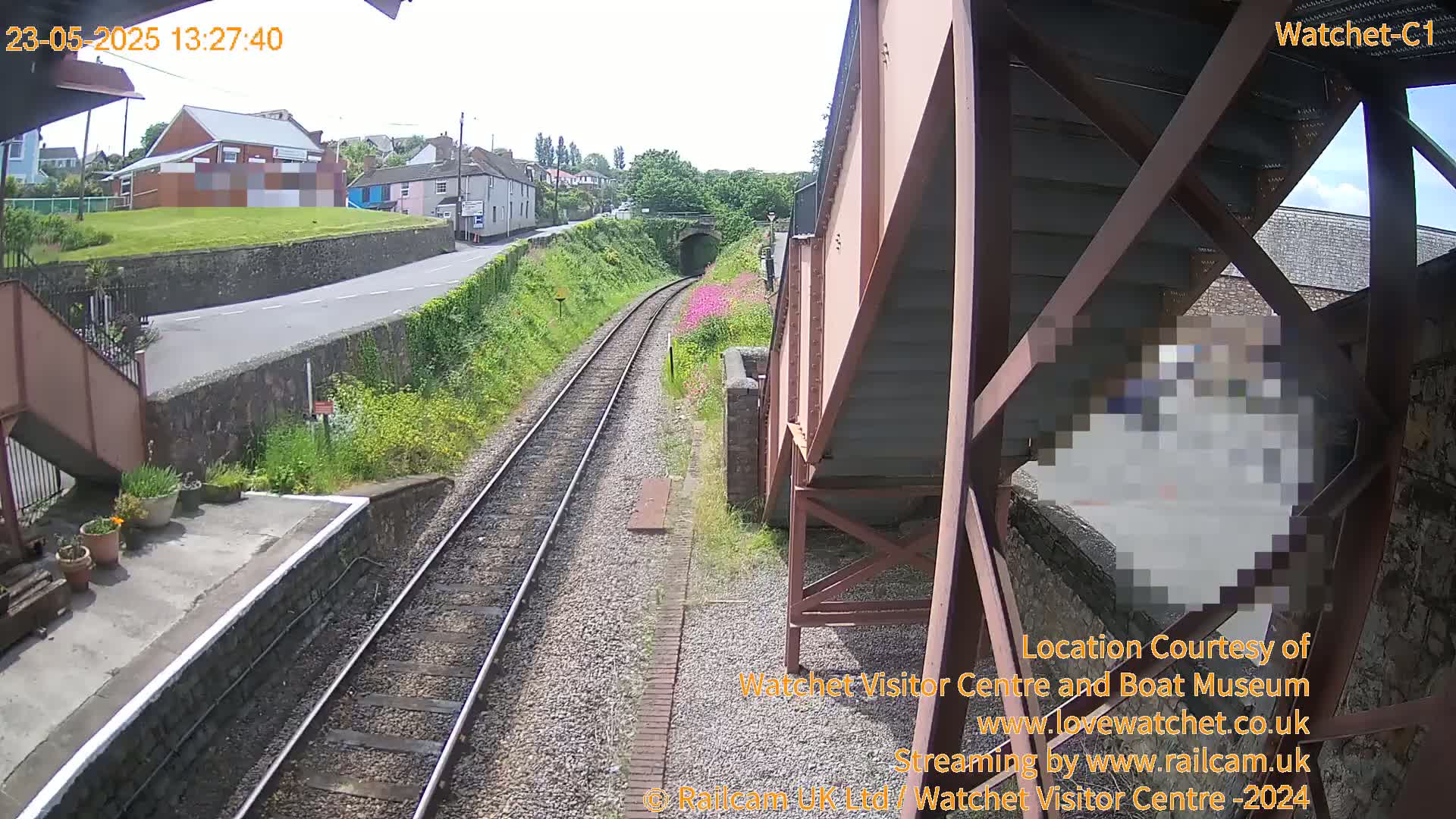 A view from a railway footbridge on a sunny day shows train tracks leading to a tunnel, a road, and buildings beyond.