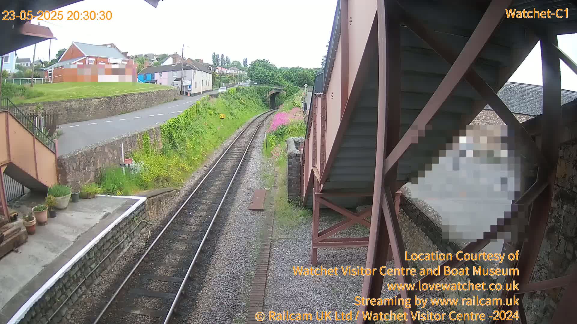 A view from a railway footbridge shows train tracks leading to a tunnel, a road, and buildings in the distance under an overcast sky.