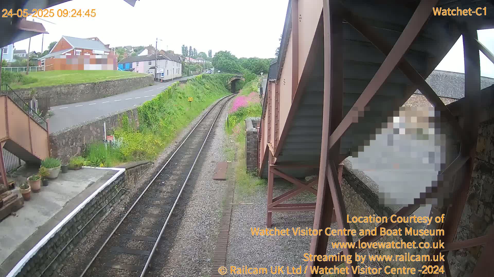 A view from a railway platform shows train tracks leading to a tunnel under a cloudy sky, with buildings and a road visible in the distance.