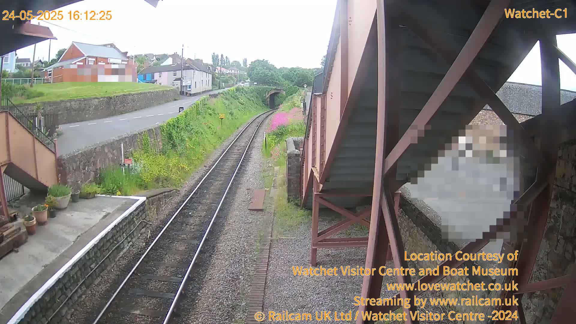 A view from a railway bridge on a partly sunny day shows train tracks leading toward a tunnel, with a road and buildings visible in the distance.