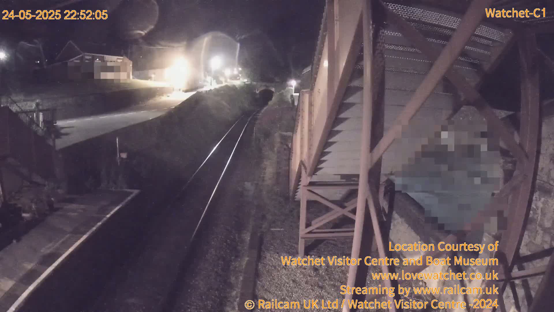 A nighttime view from a railway bridge shows train tracks, a road, and a building in the distance under dark, clear skies.