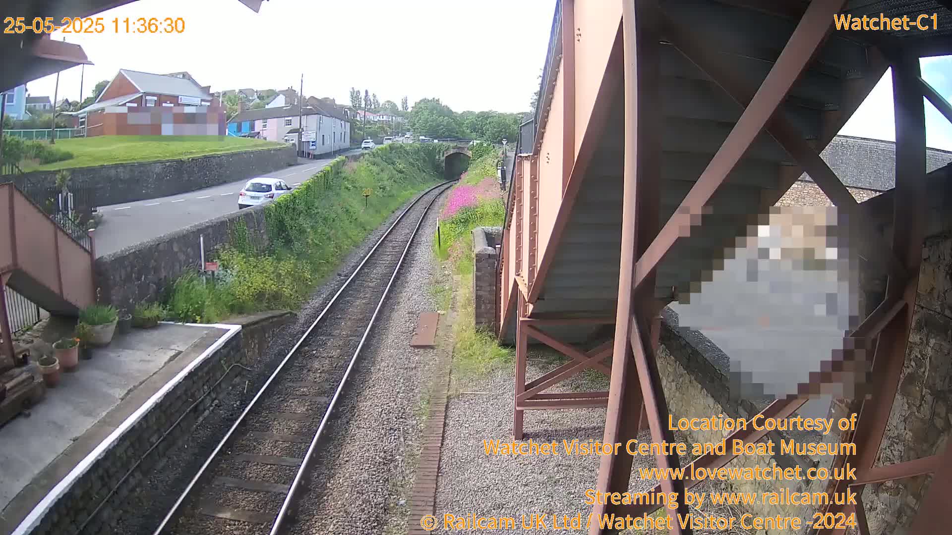 A view from a railway bridge on a sunny day shows train tracks leading to a tunnel, a road with cars, and a small town in the background.