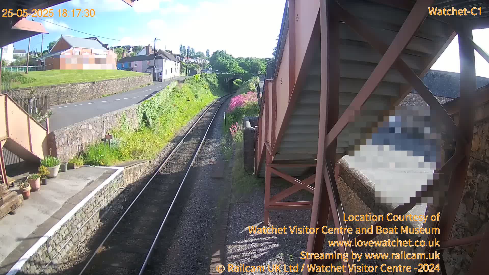 A view from a railway footbridge on a sunny day shows train tracks leading toward a small town with houses and a stone wall.