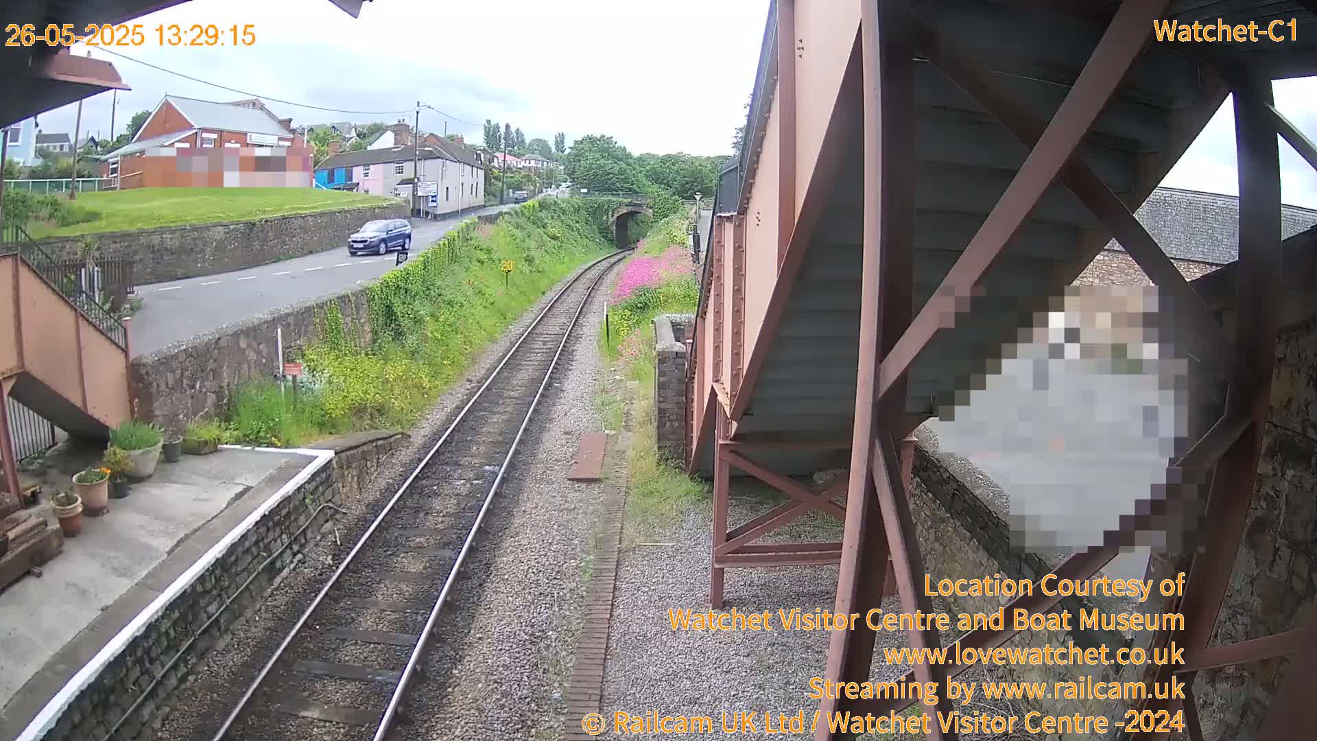 A view from a train platform shows railway tracks, a road with a car, buildings in the distance, and a partly obscured person standing under a metal structure on an overcast day.