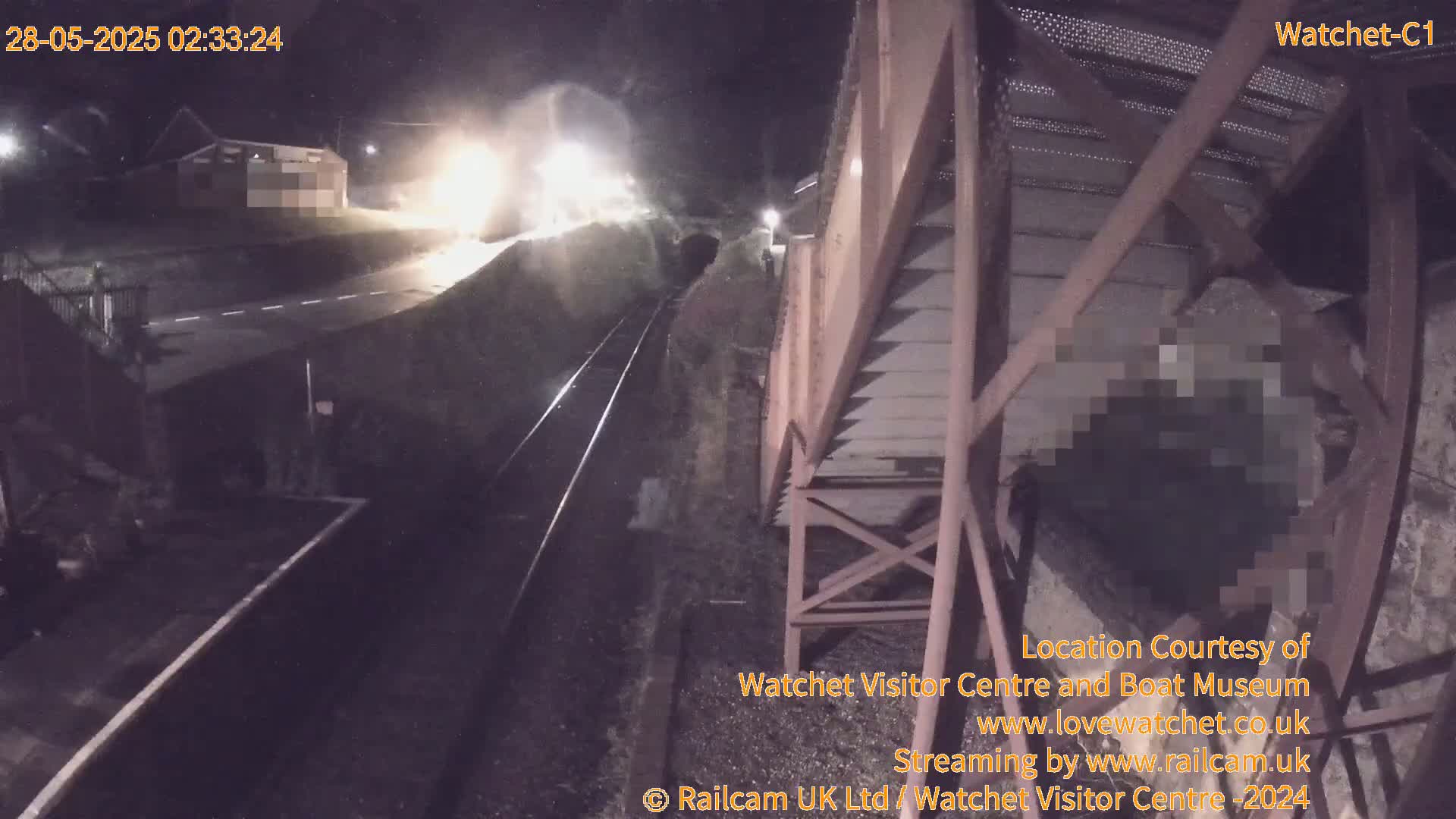 A nighttime, low-angle view shows a train passing under a metal structure next to a railway platform and road.