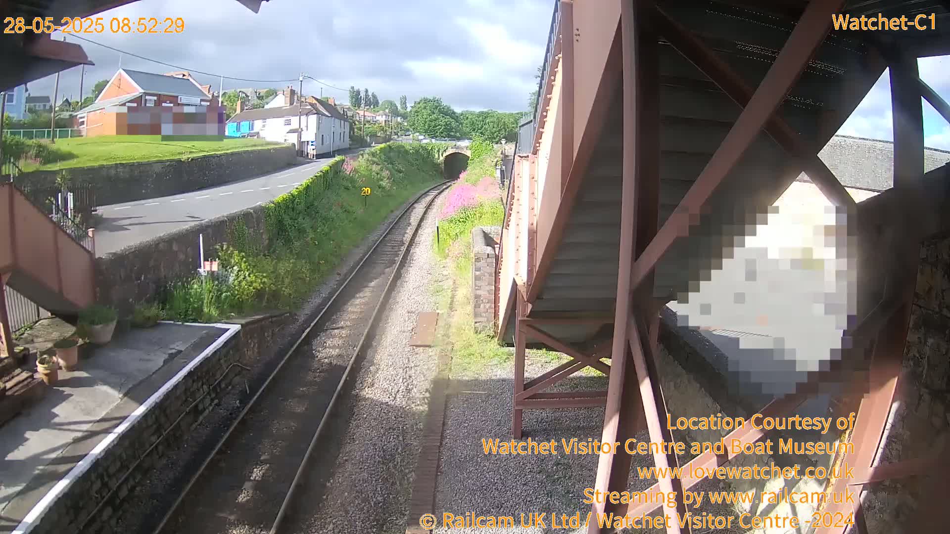A sunny view from a railway station footbridge shows train tracks leading to a tunnel, with buildings and greenery visible in the distance.