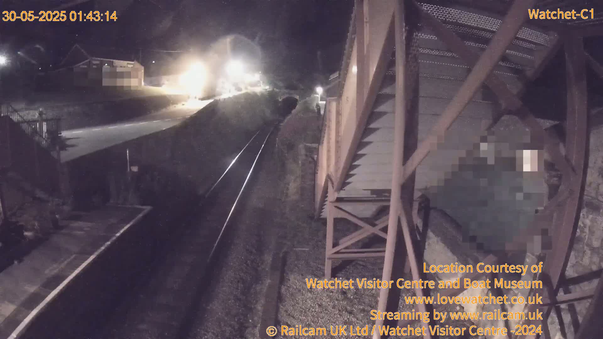 A nighttime, low-angle view shows a railway track with a train in the distance, alongside a metal structure, possibly part of a loading or unloading system.