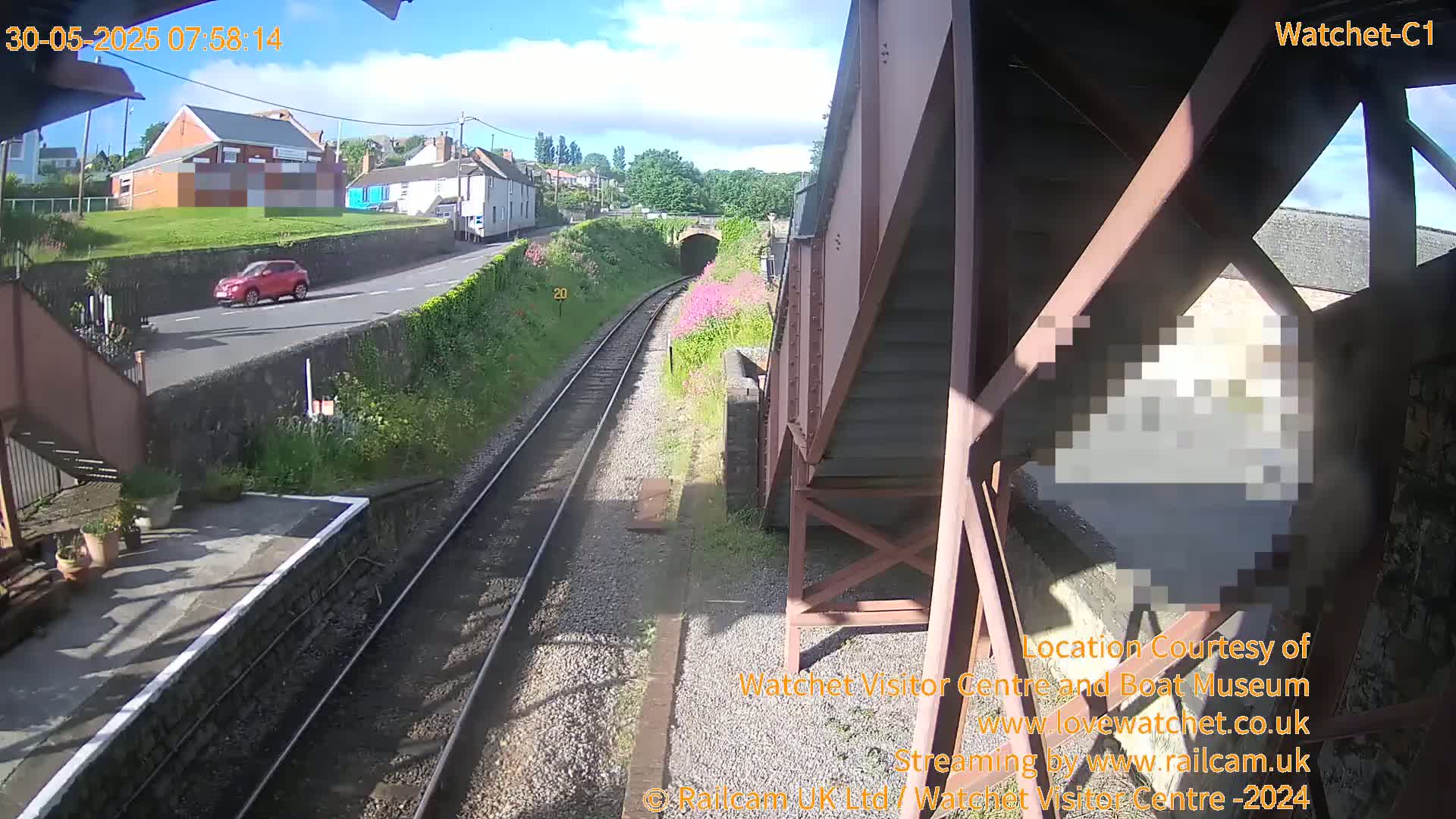 A sunny view from a railway platform shows train tracks leading to a tunnel, a red car driving on a nearby road, and a building in the distance.