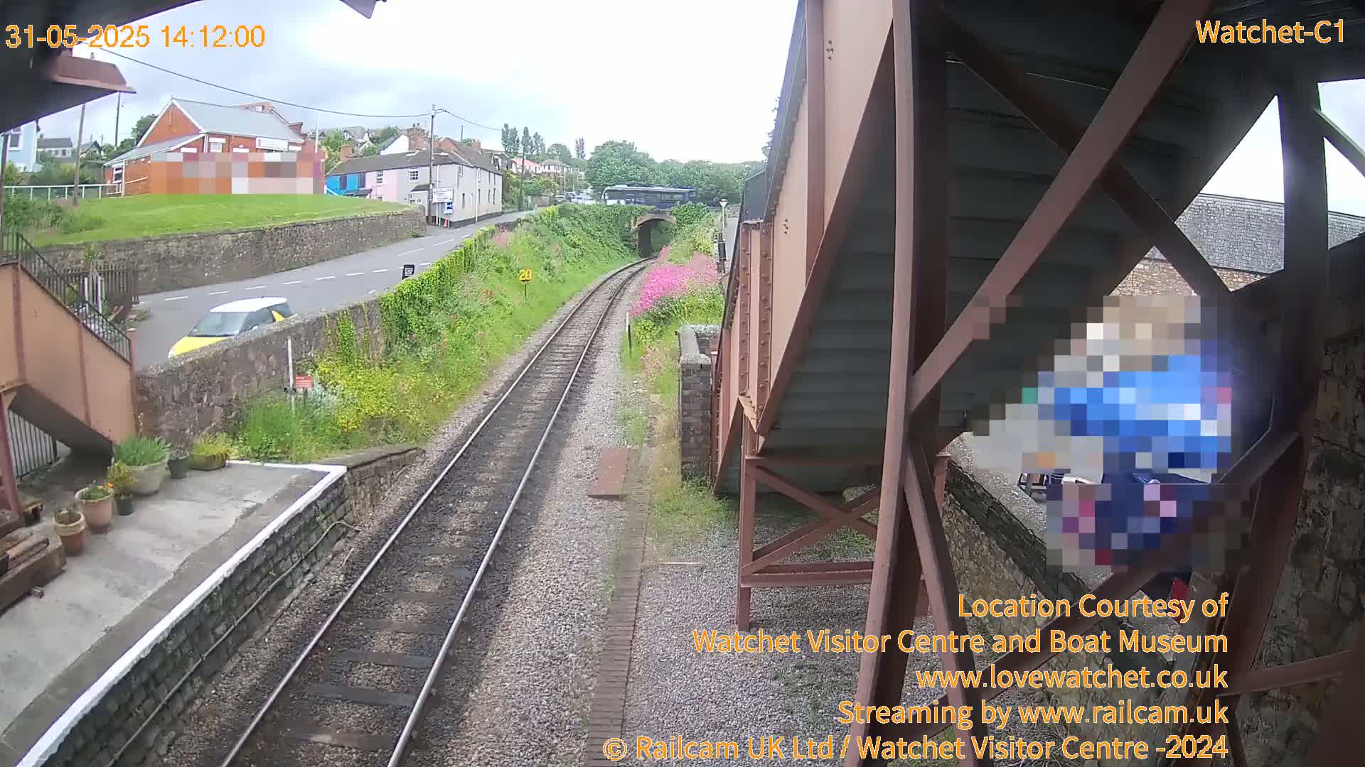 A view from a railway bridge shows train tracks, a small yellow car, and a person partially obscured by a structure under the bridge, on an overcast day.
